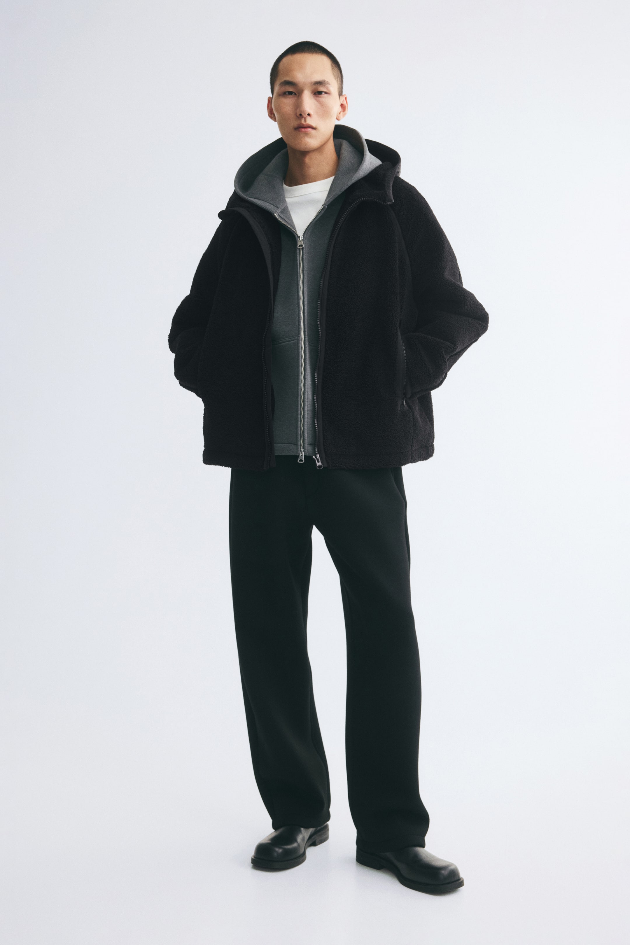 Agrandir l'image: Une veste teddy noire à capuche est portée ouverte par le mannequin, qui se tient les mains dans les poches latérales zippées. La veste est superposée à un hoodie gris zippé et à un t-shirt blanc, et est associée à un pantalon noir.