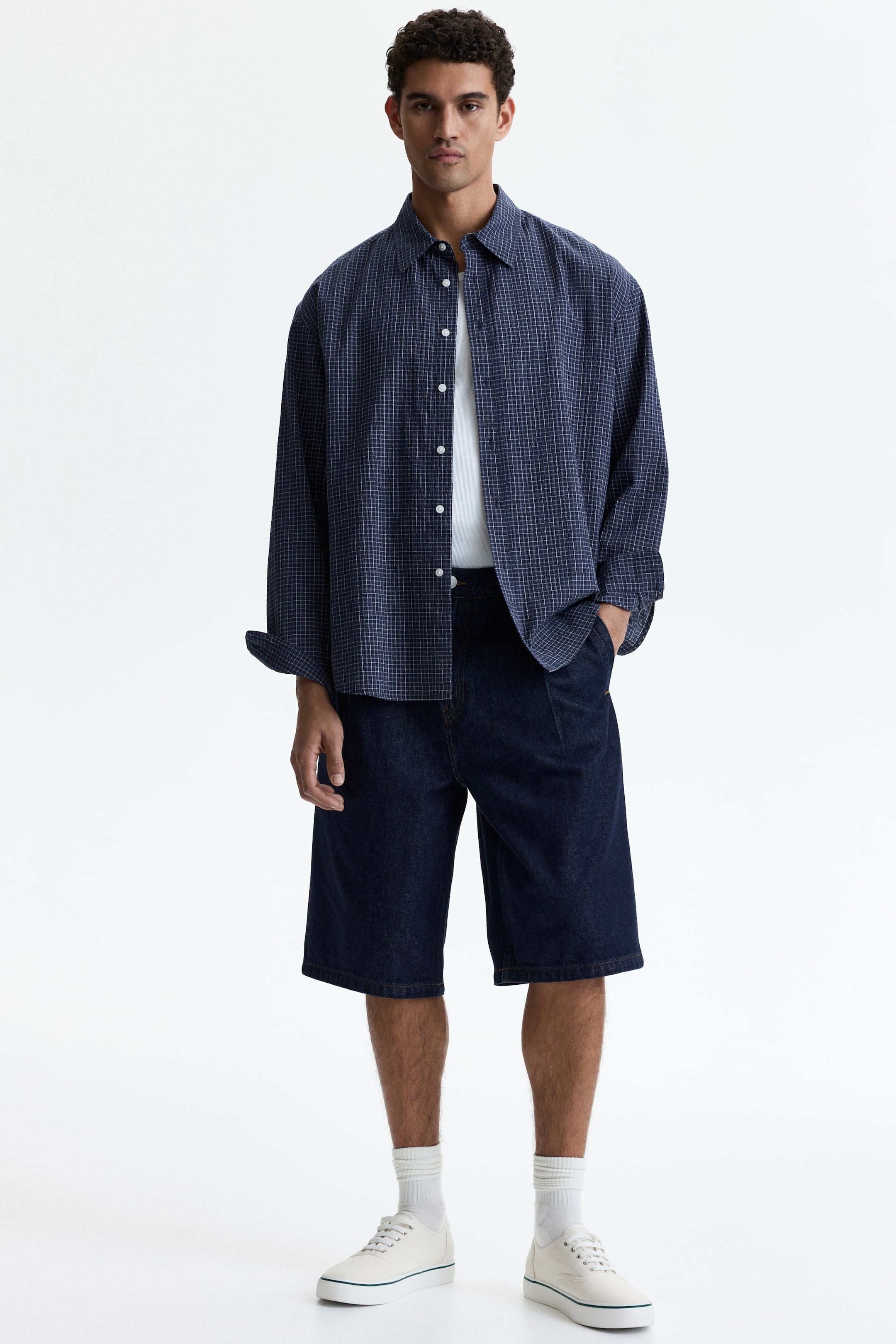 View larger image: A model is facing forward, wearing an oversized navy blue long-sleeved shirt with a small white grid pattern, worn open over a white crew-neck t-shirt. He is also wearing dark blue wide-leg denim shorts featuring front pleats, and white ribbed socks with white canvas sneakers that show a green stripe on the sole.
