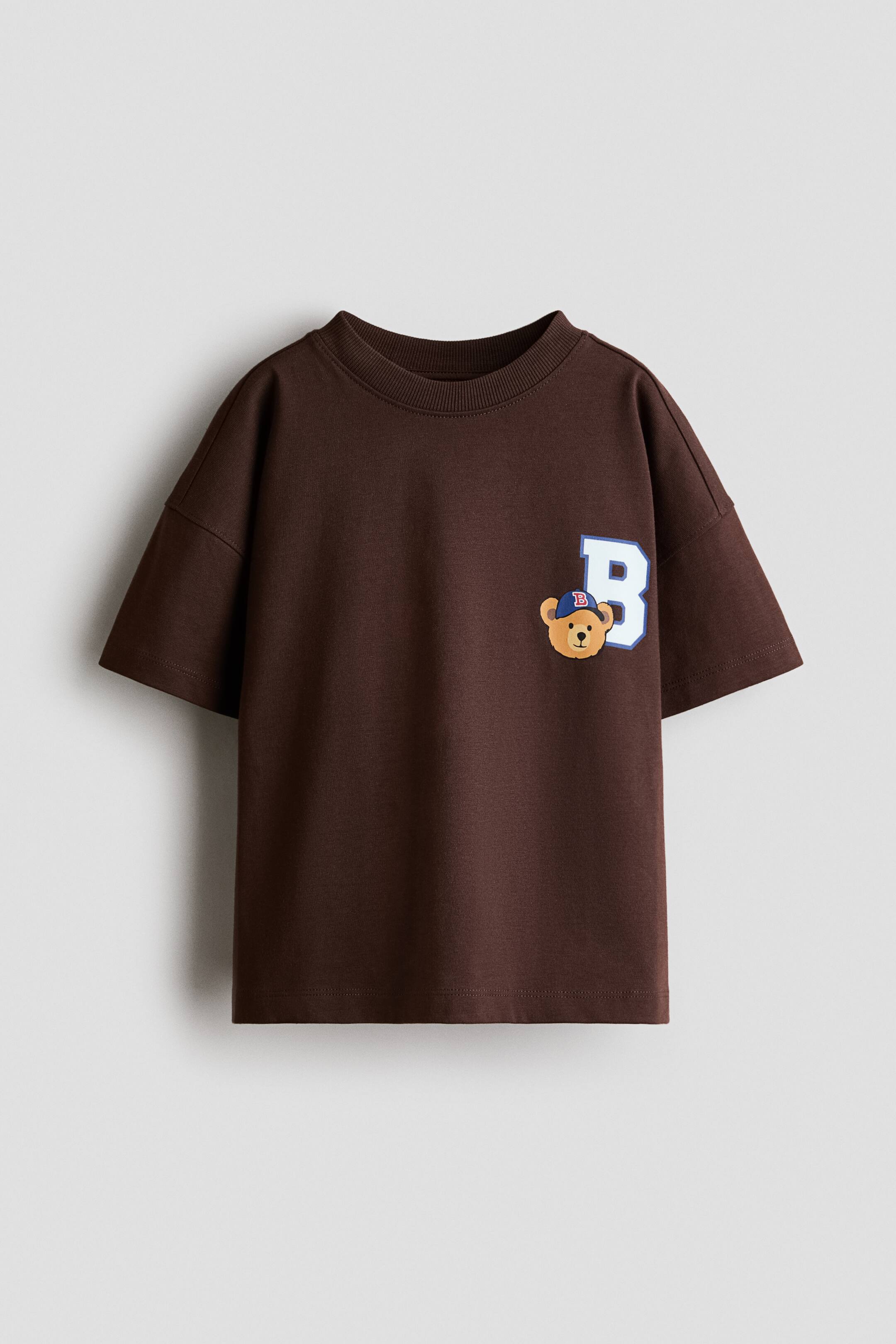 查看大圖: 一件深棕色 T 恤正面展示,採用羅紋圓領、落肩設計與短袖。左胸位置印有一個大型白色字母「B」,外框以藍色勾勒,並有一隻戴著藍色棒球帽的卡通泰迪熊重疊於圖案上。