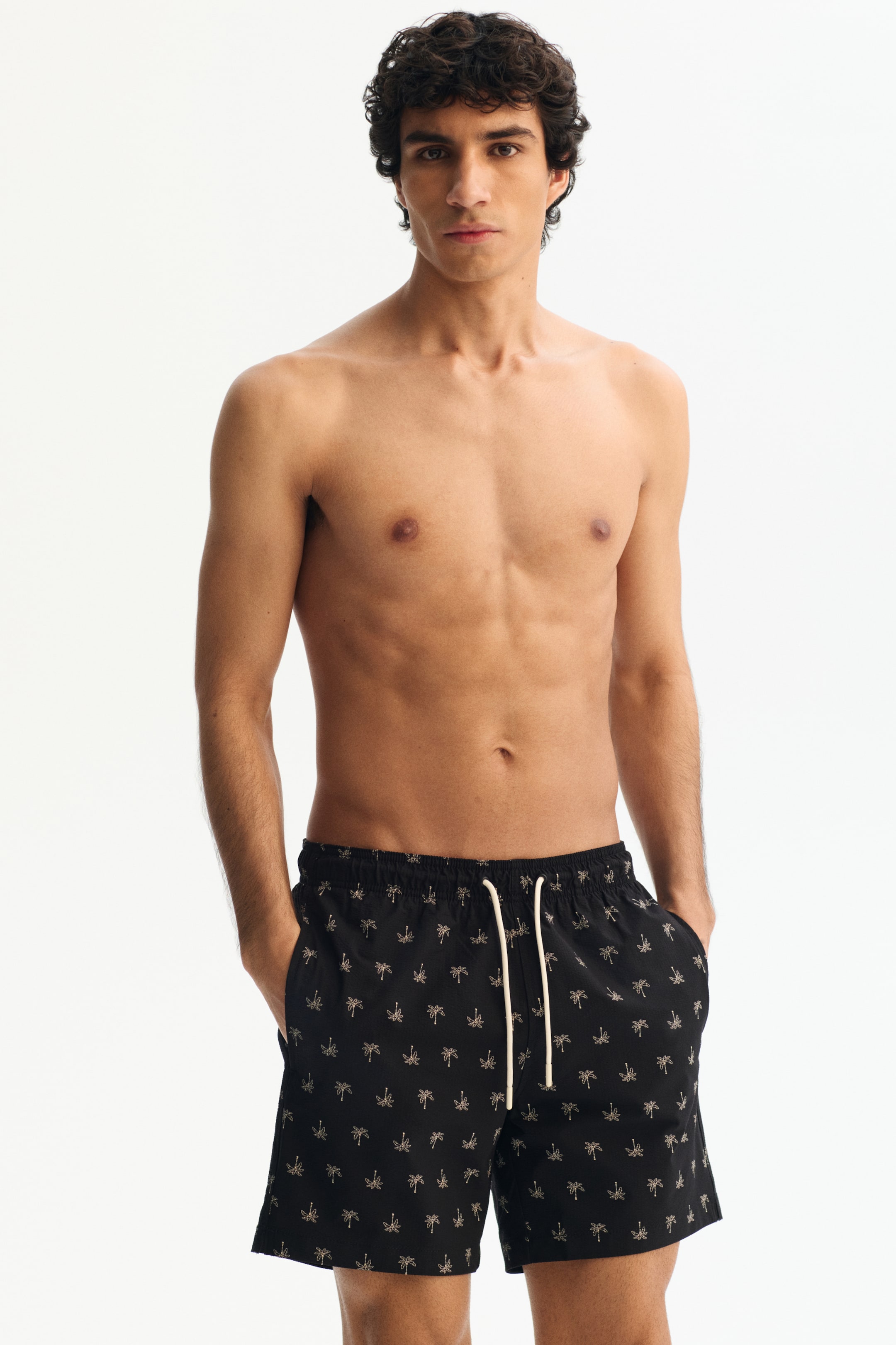 이미지 확대 보기: 전면을 향해 있는 검은색 수영 반바지는 연한 색상의 야자수 패턴이 반복되어 있습니다. 이 반바지는 신축성 있는 허리밴드와 앞쪽에 연한 색상의 드로스트링, 그리고 옆주머니가 디자인되어 있으며, 느슨하게 착용되며 허벅지 중간까지 내려옵니다.