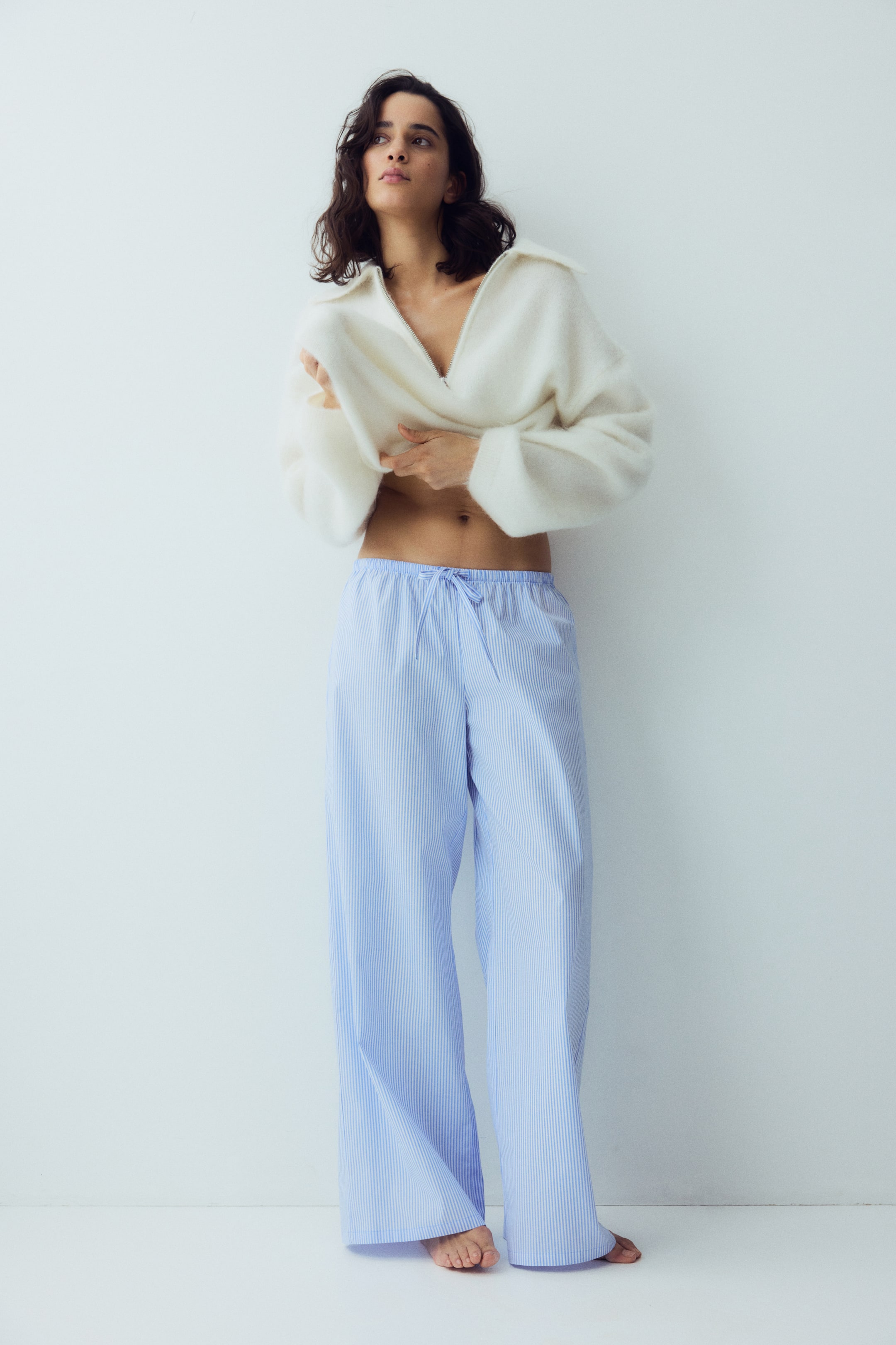 Xem ảnh lớn hơn: Một chiếc áo len dệt kim trắng mịn với khóa kéo nửa chừng và cổ áo rộng đang được kéo lên, để lộ phần bụng. Quần pajama ống rộng màu xanh nhạt, có sọc dọc màu trắng mỏng và dây rút ở eo, đang được mặc, tạo thành những nếp gấp nhẹ ở chân. Cả hai món đồ được trình bày ở mặt trước.
