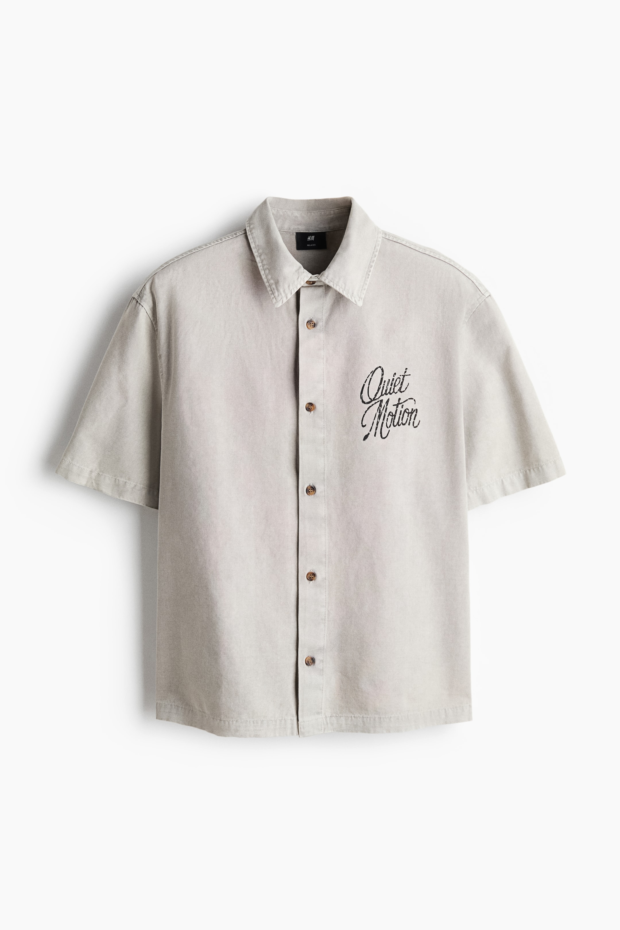 Voir une image plus grande: Une chemise à manches courtes gris clair est présentée de face, avec un col classique à larges pointes, une fermeture boutonnée complète avec boutons bruns et un texte brodé noir indiquant « Quiet Motion » sur la poitrine gauche.