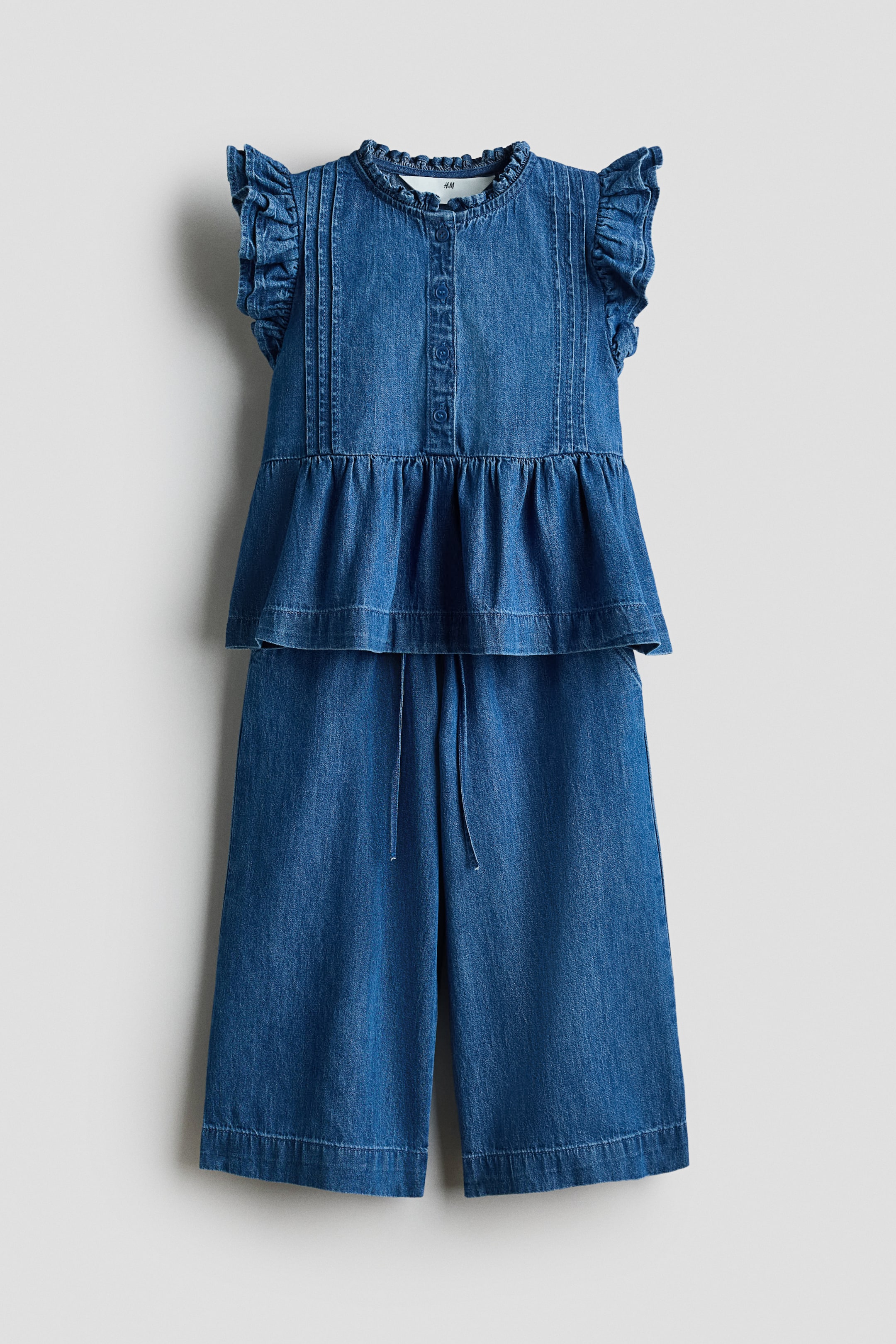 Agrandir l'image: Un ensemble en denim deux pièces est présenté, comprenant un haut bleu à basque avec un col montant à volants, des manches courtes à volants, une patte de boutonnage sur le devant, des plis nervurés verticaux sur le corsage et un ourlet à basque froncé. Un pantalon large assorti complète l'ensemble, avec une taille élastiquée à cordon de serrage et des jambes raccourcies.