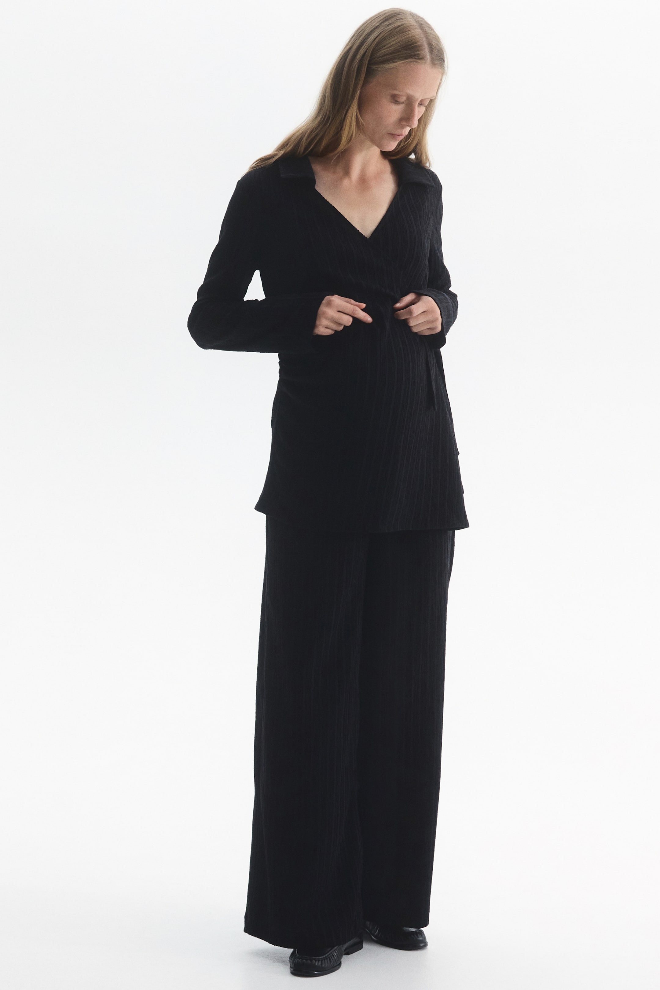 View larger image: A black textured jersey set, featuring a long-sleeve wrap top with a collar and matching wide-leg pants, is being worn by the model. The model is adjusting the tie closure at the side of the wrap top.