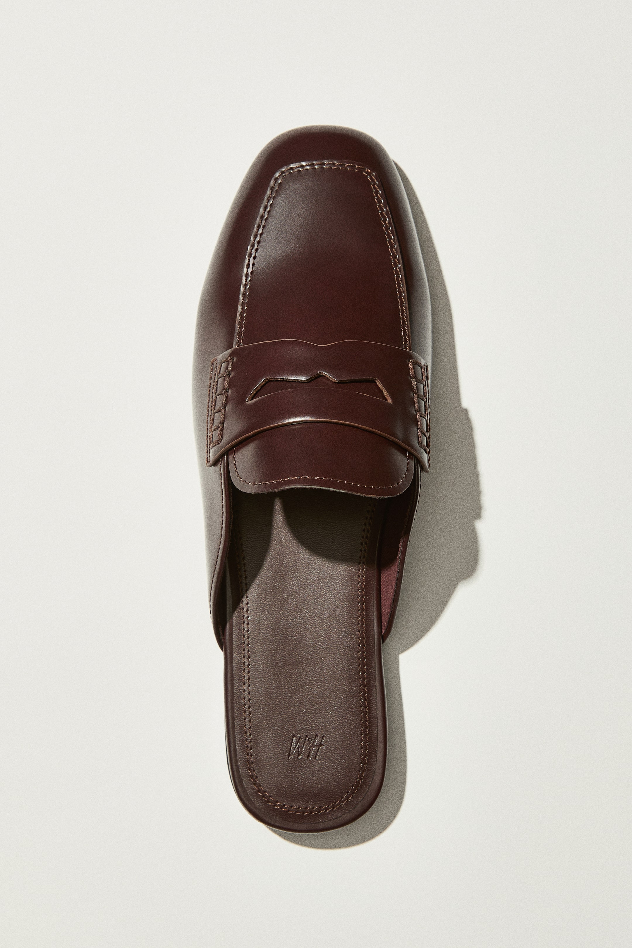 View larger image: A dark brown loafer mule is showcased from a top-down perspective, highlighting an almond toe and a penny keeper strap across the vamp. The sleek upper features visible stitching around the toe box, and an H&M logo is embossed on the footbed.