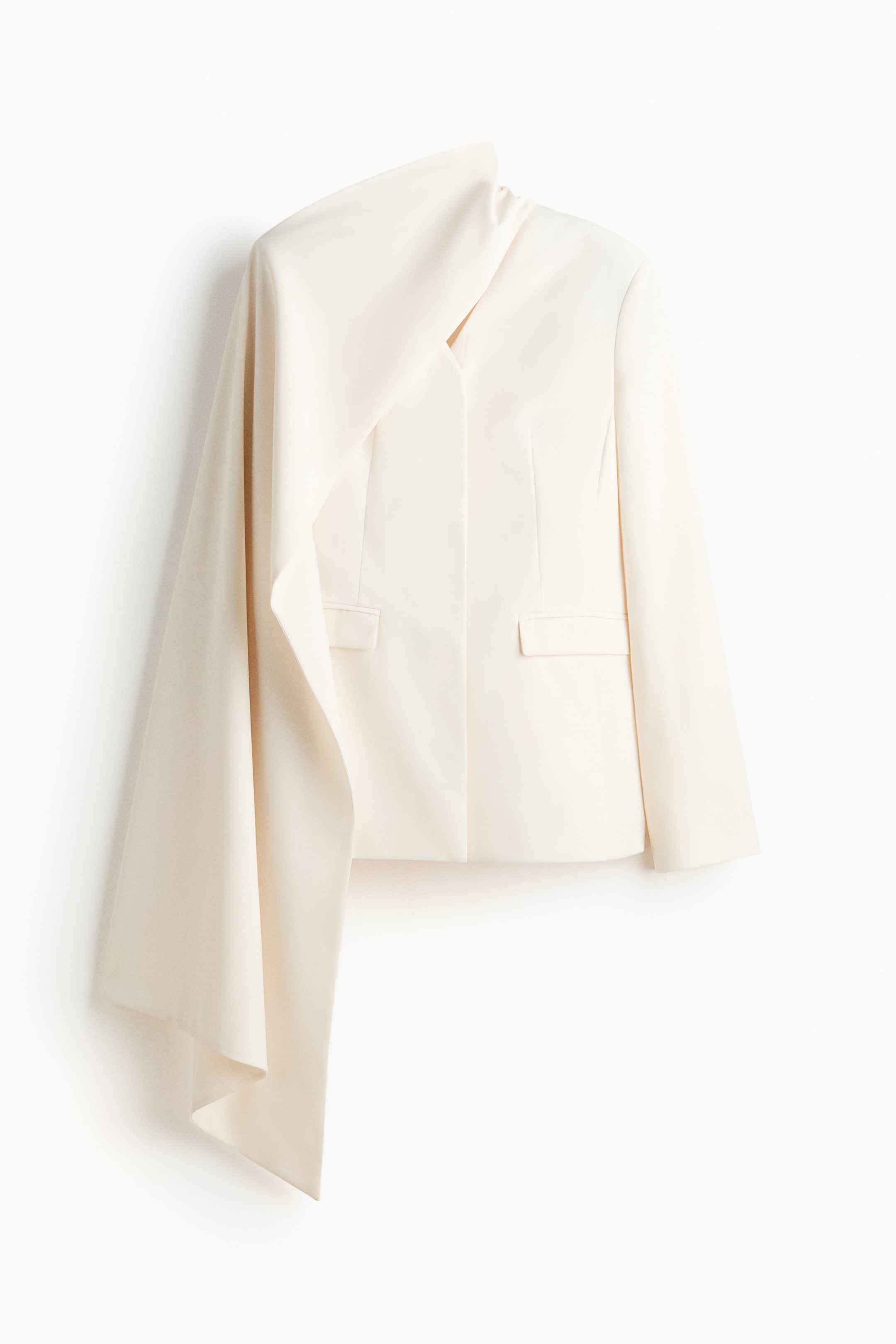 View larger image: An off-white blazer is presenting its front, featuring a structured body with two flap pockets and an asymmetrical design. A large, draped fabric panel is extending from the left shoulder, flowing down the side of the blazer.