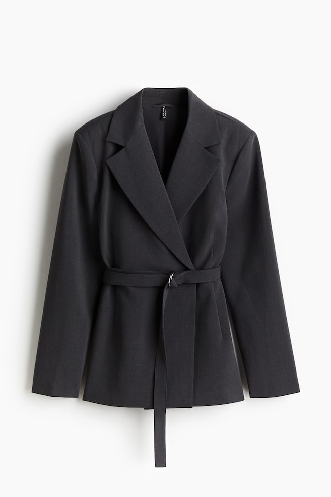 A dark grey blazer is being shown from the front, featuring wide notch lapels and a wrap-front design. A matching self-fabric belt is cinching the waist, with one end being secured by a silver D-ring buckle and the other end hanging loose. - 1