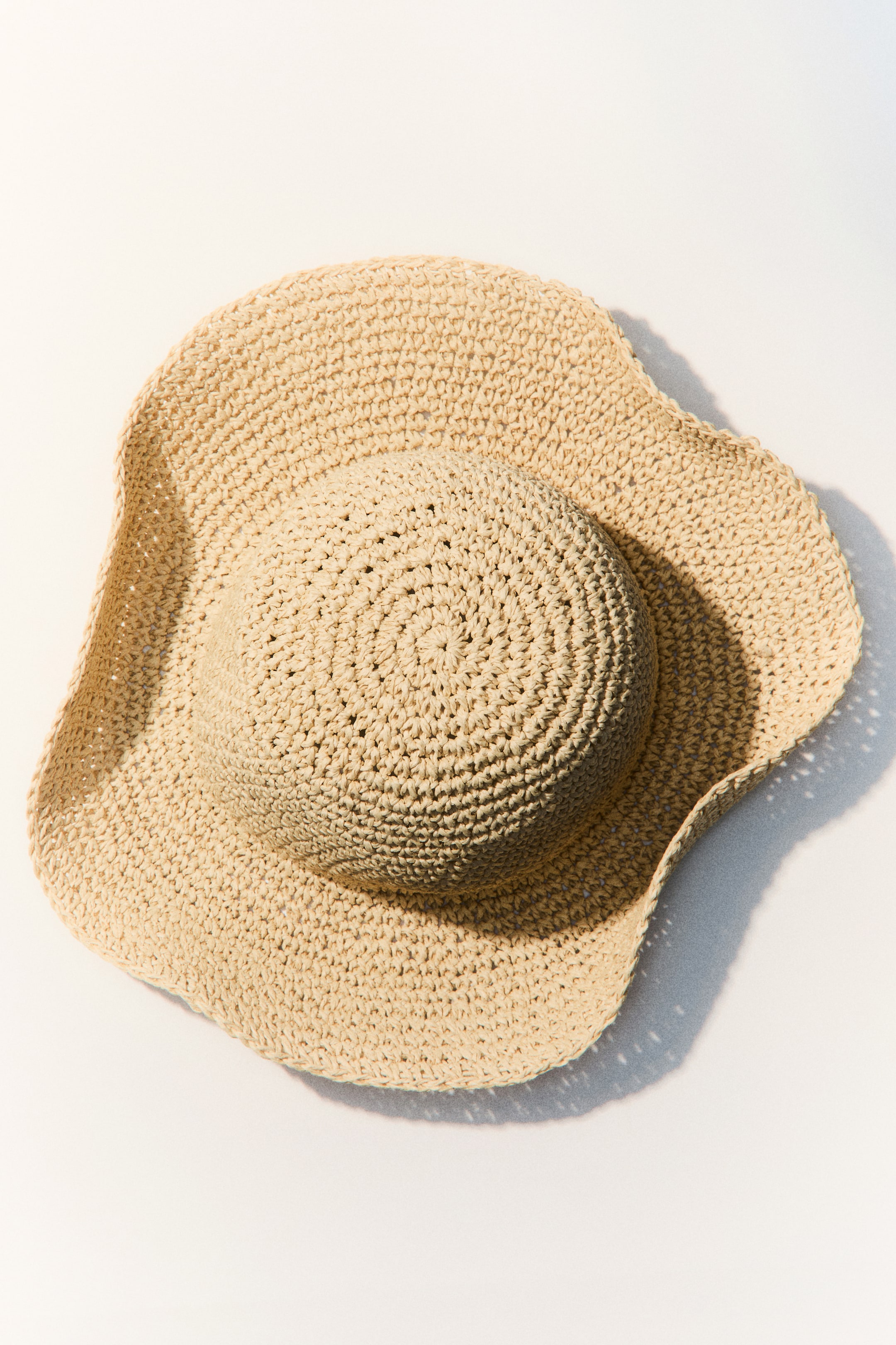 View larger image: A light beige crocheted hat is lying flat, showcasing its round crown with concentric stitching and a wide, wavy brim.