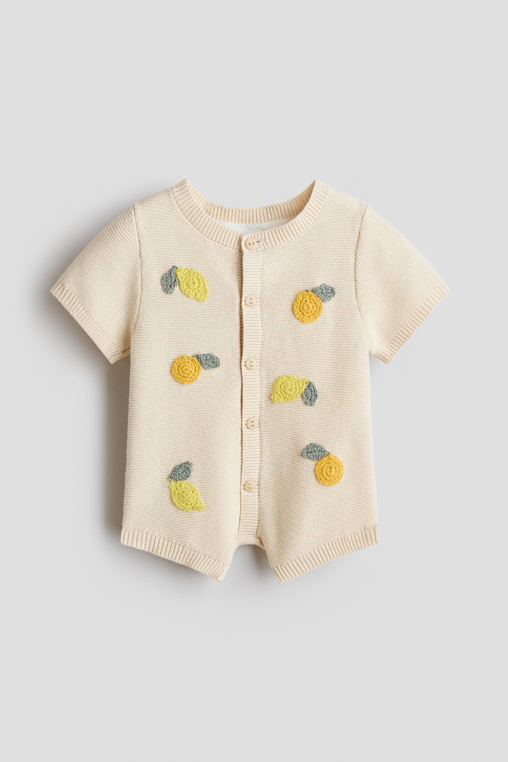 Ver imagem maior: Um babygrow de malha em creme apresenta uma carcela de botões frontal ao longo de todo o comprimento, mangas curtas e uma gola redonda canelada. Está adornado com motivos de limão e folhas em croché amarelos e verdes dispersos, e termina com punhos canelados nas mangas curtas e aberturas das pernas.