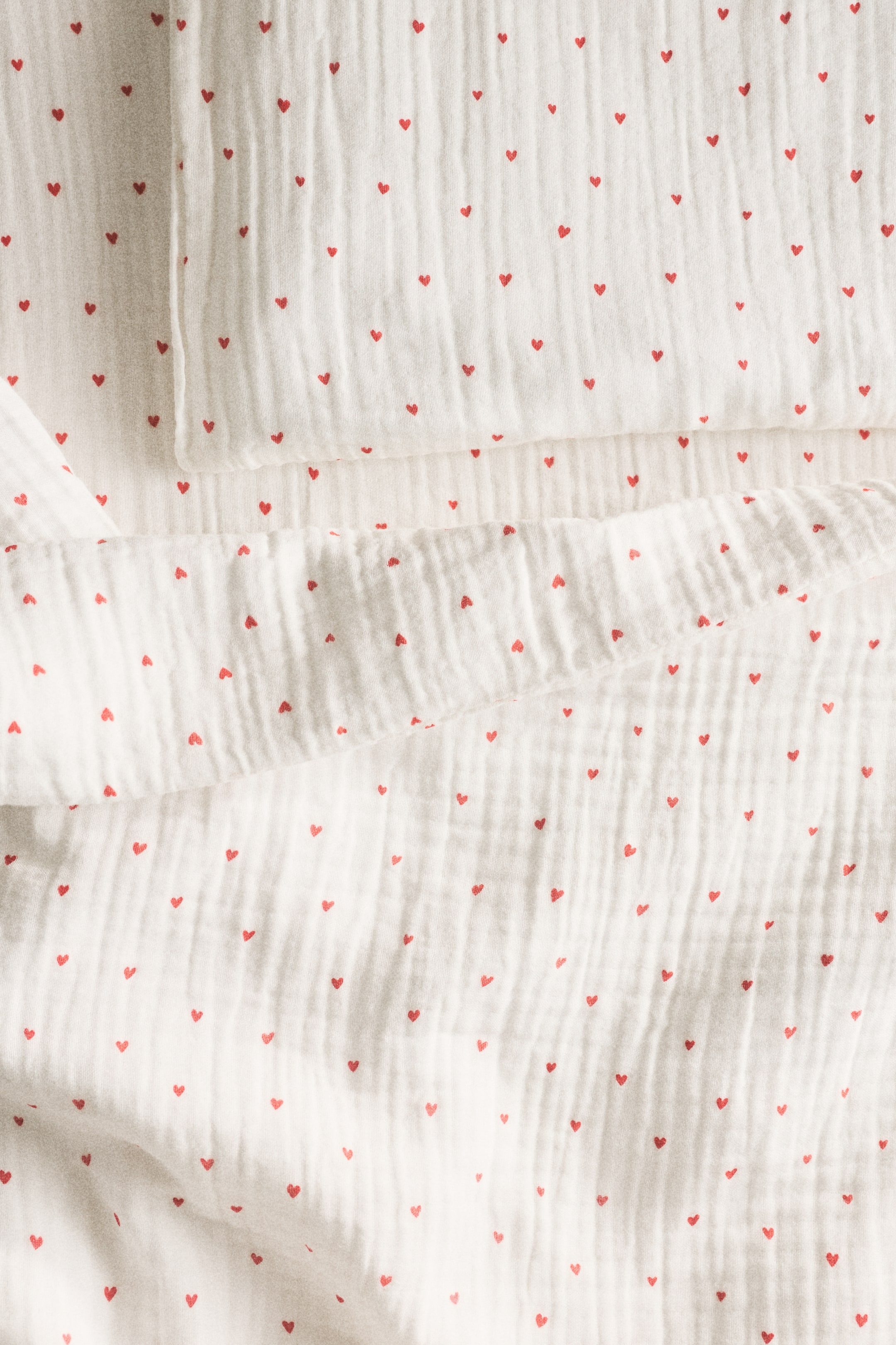 View larger image: A cot duvet cover set features a white, crinkled cotton muslin fabric adorned with small red hearts scattered across its surface. The set includes a duvet cover, neatly folded, and a matching pillowcase, elegantly layered at the top left.