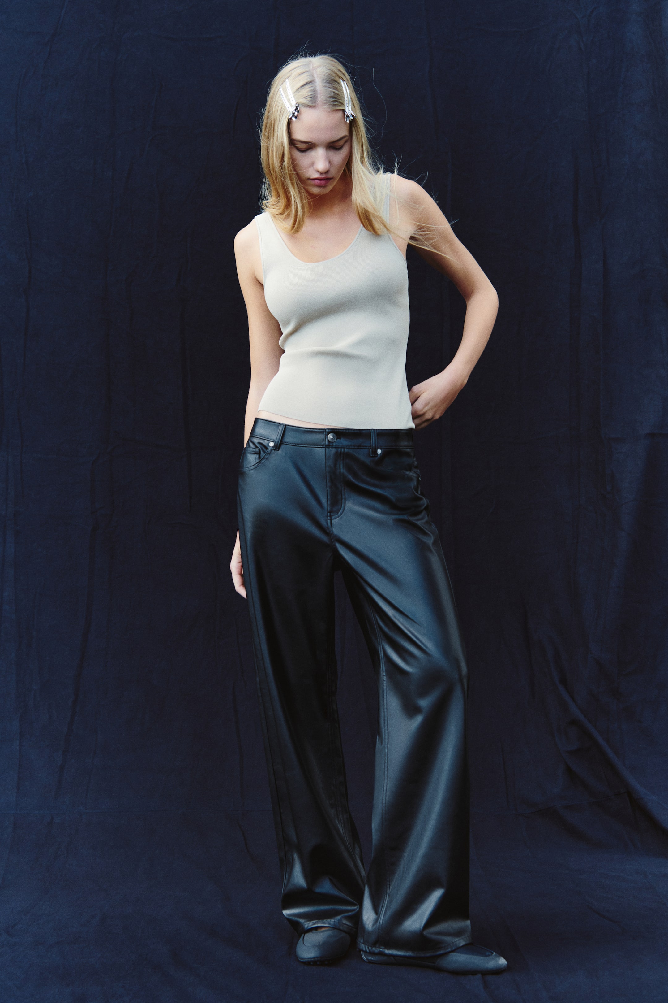 Grotere afbeelding bekijken: De model draagt een baggy broek met vijf zakken in zwarte gecoate stof, met een wijde pijp en een zichtbare tailleband met knoopsluiting. De broek is gestyled met een lichtbeige geribde tanktop en donkere platte schoenen.