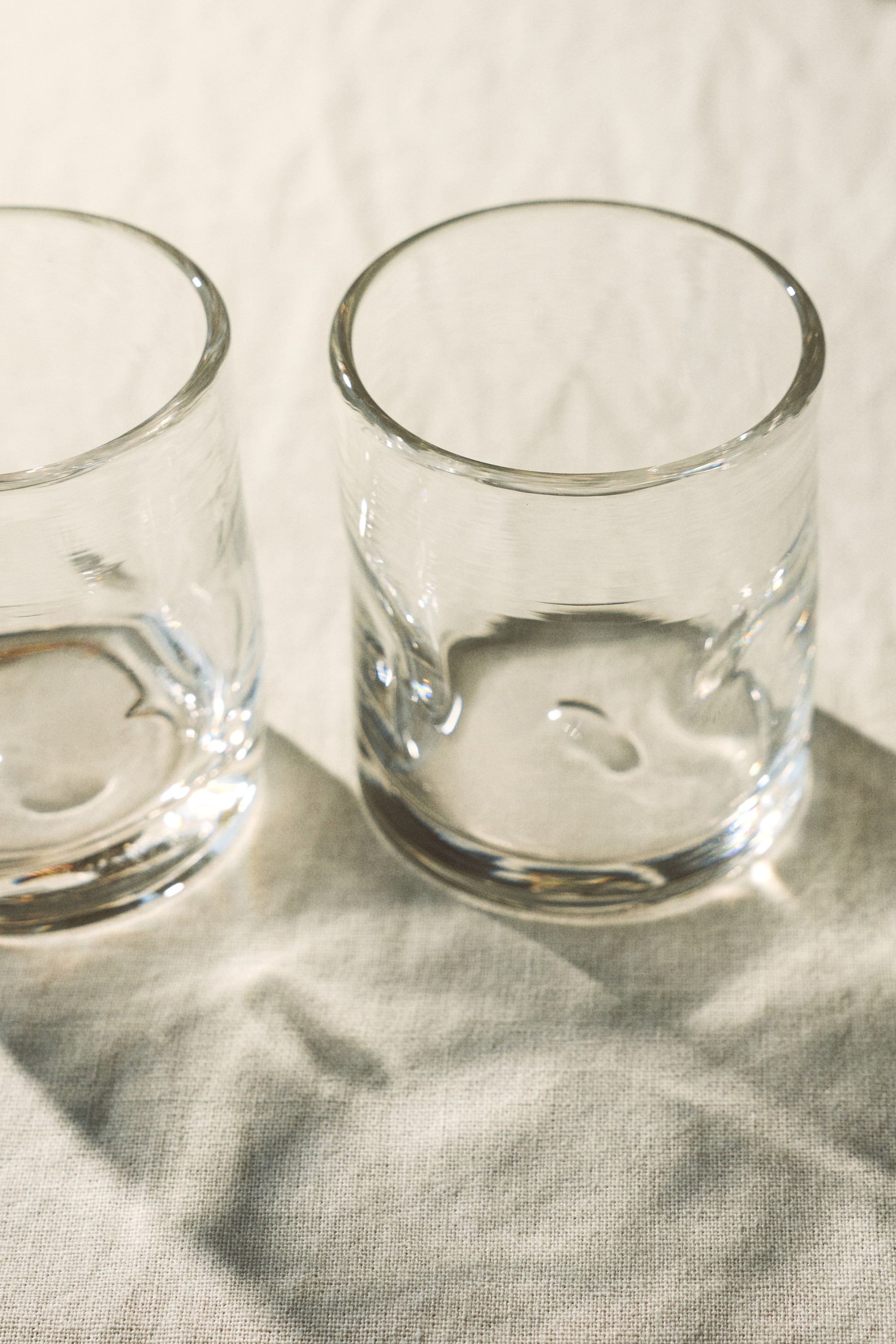 View larger image: Two clear drinking glasses are positioned side-by-side, each showcasing subtly irregular, wavy textures along their sides and clear, rounded rims. The prominent right glass is angled towards the left, highlighting its distinctive shape.