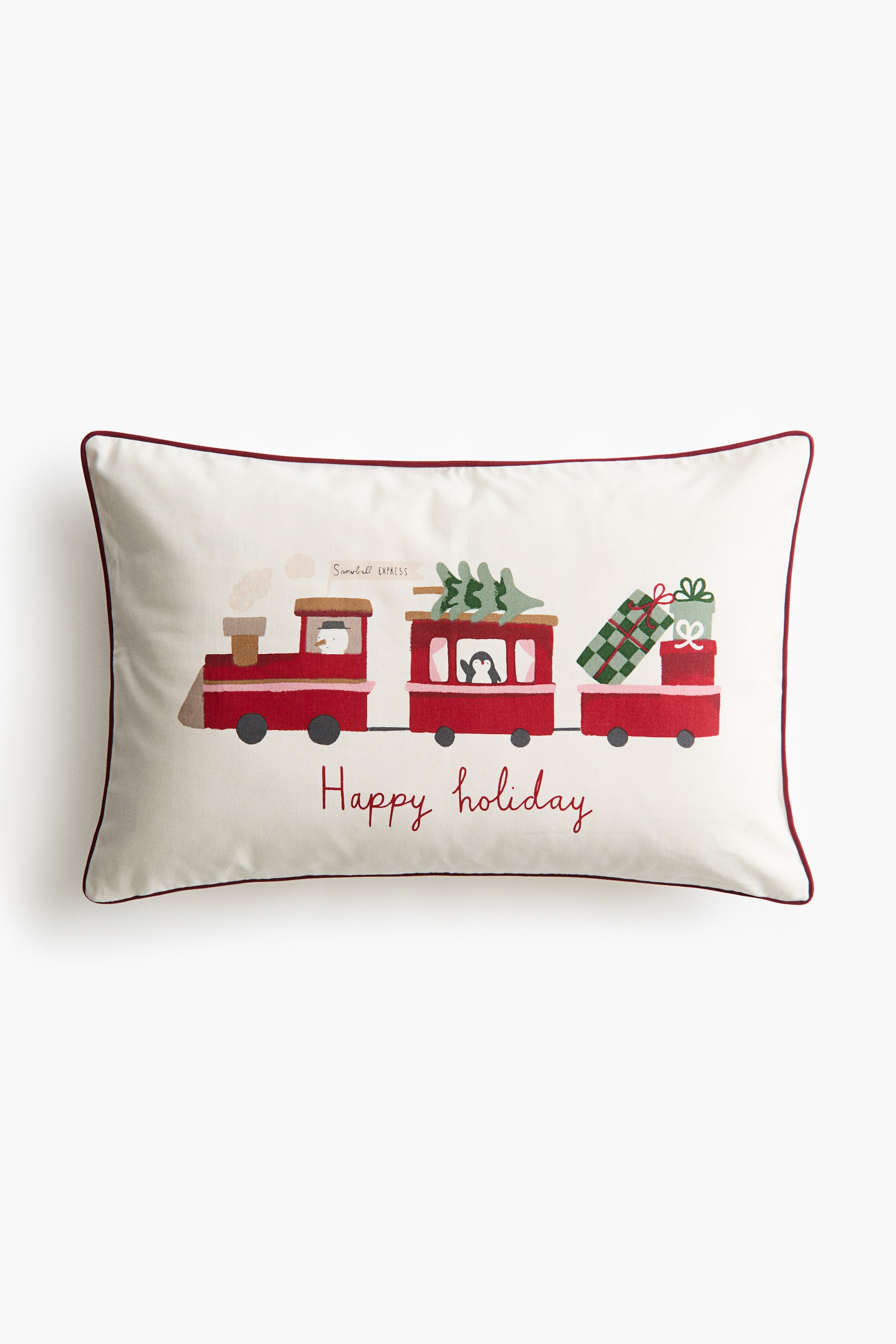 View larger image: A rectangular off-white throw pillow is shown front-facing, featuring a slim red piped border and a central illustration of a red three-car holiday train. The train is carrying a snowman, a penguin, a green tree, and wrapped gifts, with the red cursive text "Happy holiday" written below.
