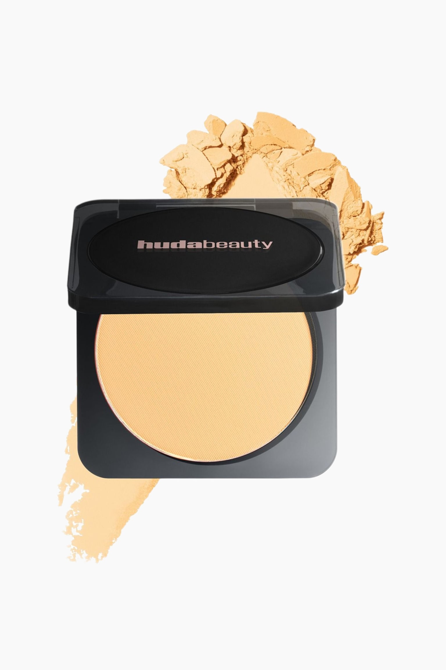 Musta Huda Beauty -pressattu puuterikompakti on avattuna, ja siinä näkyy pyöreä vaaleankeltainen puuteripannu. Irtopuuteri sirottautuu kompakista taaksepäin ja vasemmalle, ja puuterinäyte näkyy alapuolella.