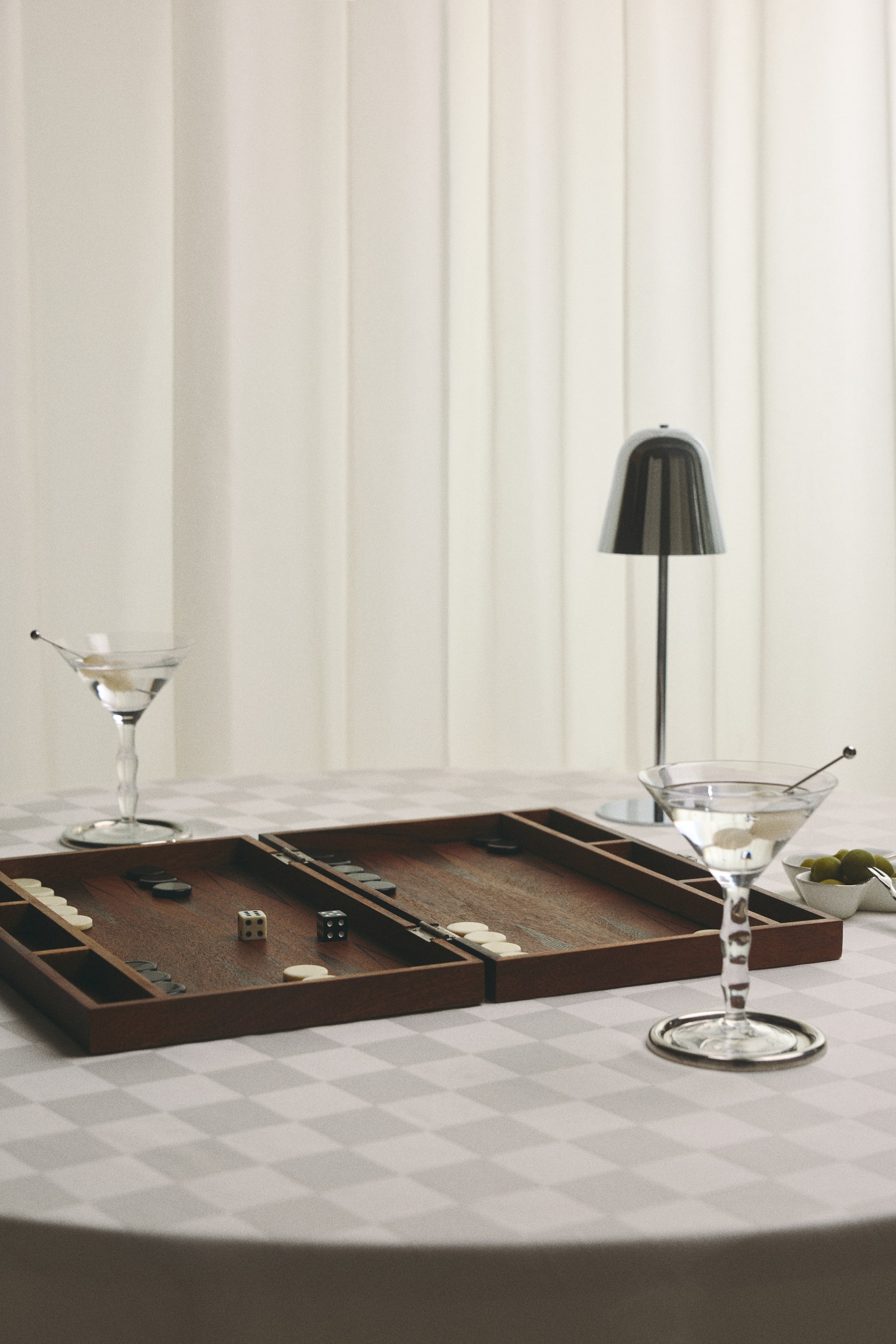 View larger image: A classic wooden backgammon set is lying open, showcasing its two hinged rectangular trays with a rich brown wood grain. The board displays round cream and black checkers positioned on the playing field, alongside one white and one black die resting near the centre.