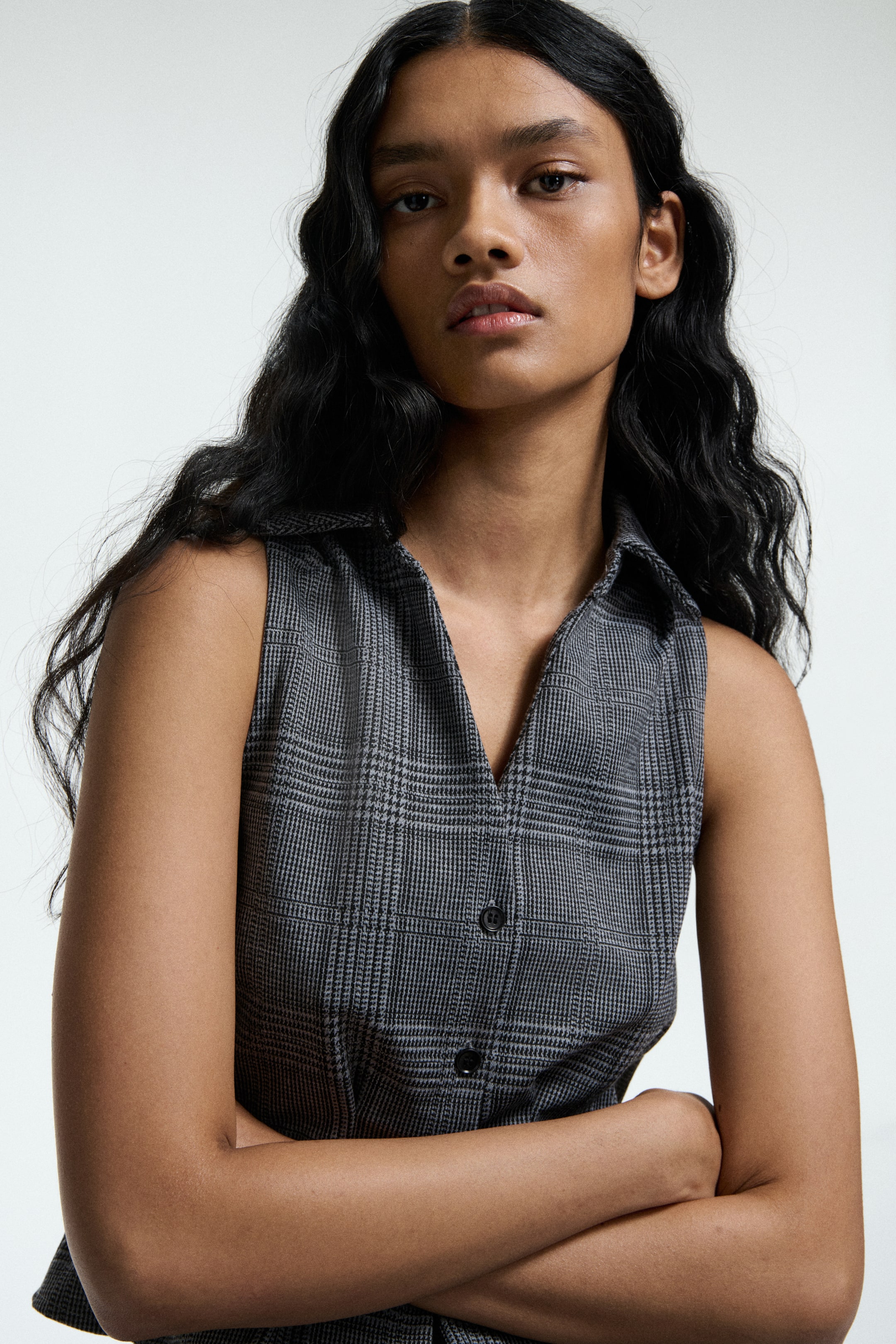 View larger image: A sleeveless top in a grey Prince of Wales check pattern is being presented from a slight front-right angle. It is featuring a pointed collar, a V-neckline, and a button-front closure with two visible black buttons.