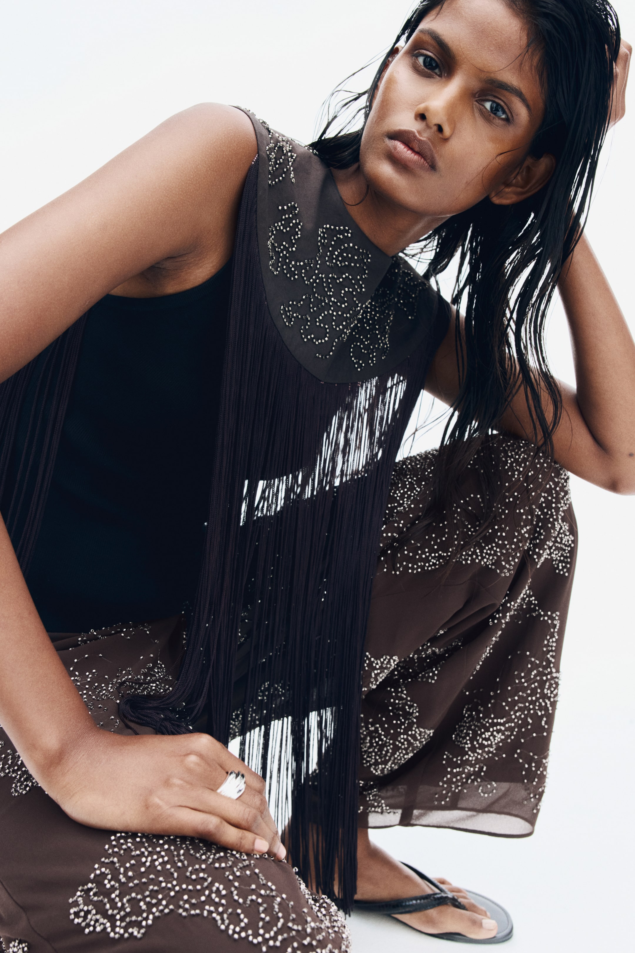 View larger image: Loose-fit brown pants in a sheer, lightweight fabric embellished with patterns of sparkling silver beads are being worn by the model crouching down. The pants are paired with a black sleeveless top, a brown beaded collar with long cascading fringe, and black flat sandals.