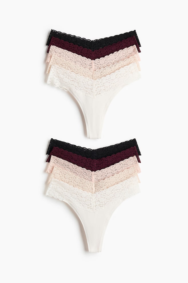 Zwei identische Sets aus fünf Tanga-Slips liegen flach, jedes Set zeigt einzelne Tangas in den Farben Off-White, Hellbeige, Hellrosa, Burgunderrot und Schwarz, alle mit einer Spitzenverzierung entlang der Taille. - 1