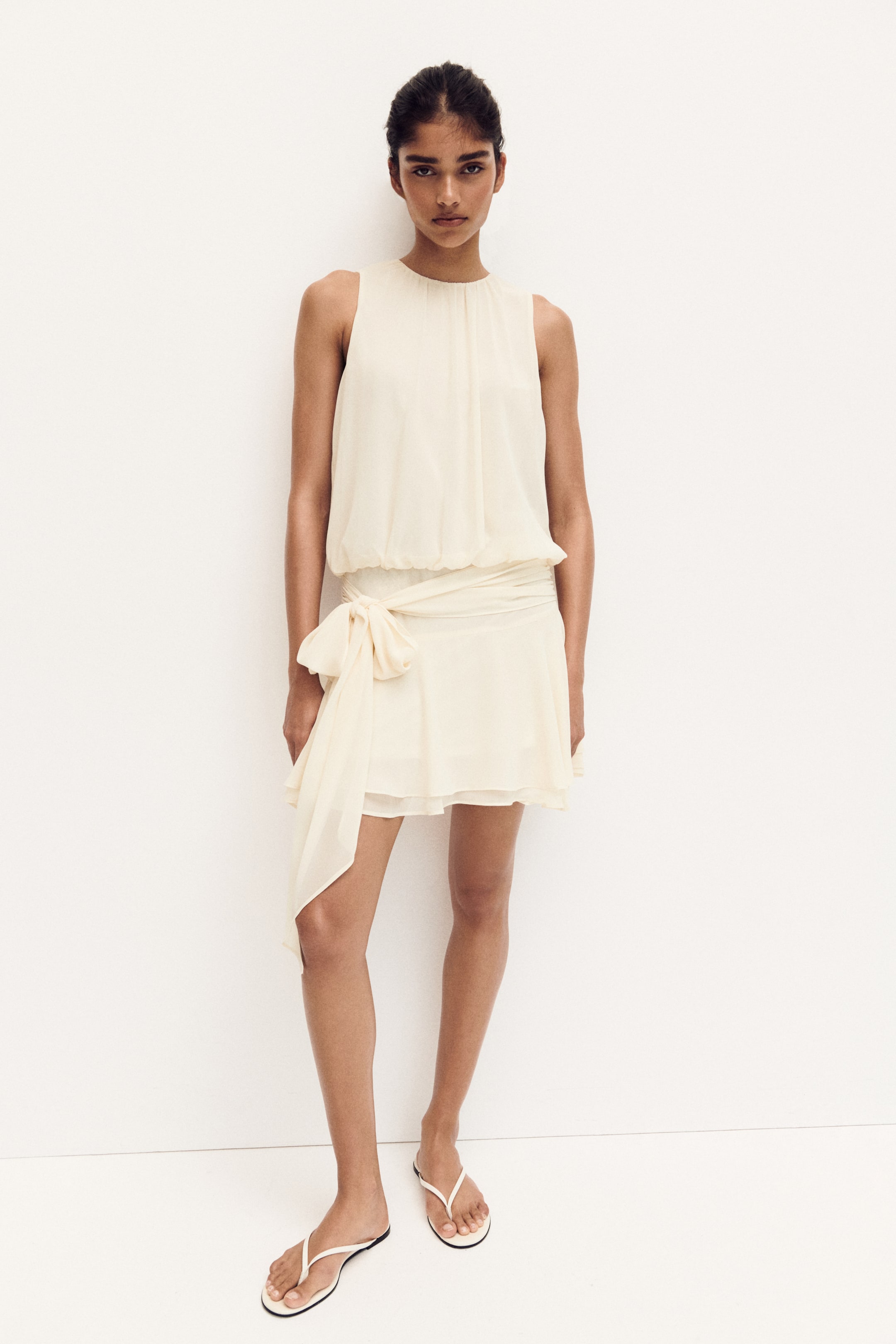 View larger image: The light cream sleeveless dress showcases a gathered round neckline and a blouson top that drapes gracefully. Its elasticized waist is accentuated by a wide sash, tied into a bow on the left hip, cascading into a short, layered skirt.