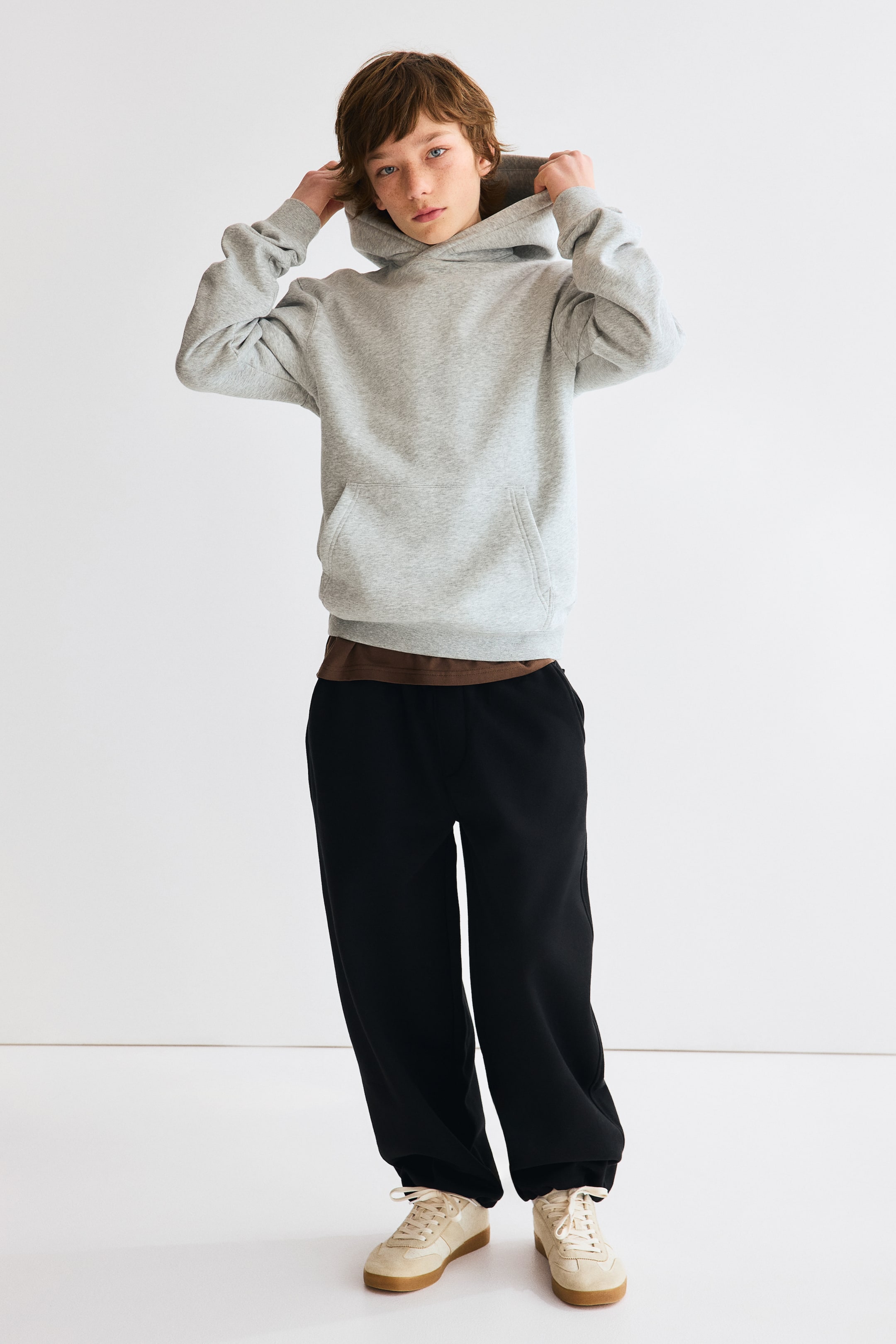 Visualizza immagine più grande: Una felpa con cappuccio grigio chiaro presenta un cappuccio, una tasca frontale a marsupio e maniche lunghe con polsini a coste, offrendo una vestibilità rilassata sopra una t-shirt marrone. I pantaloni della tuta neri hanno una vestibilità ampia e comoda, che si stringe alle caviglie, e sono abbinati a sneakers beige chiaro.