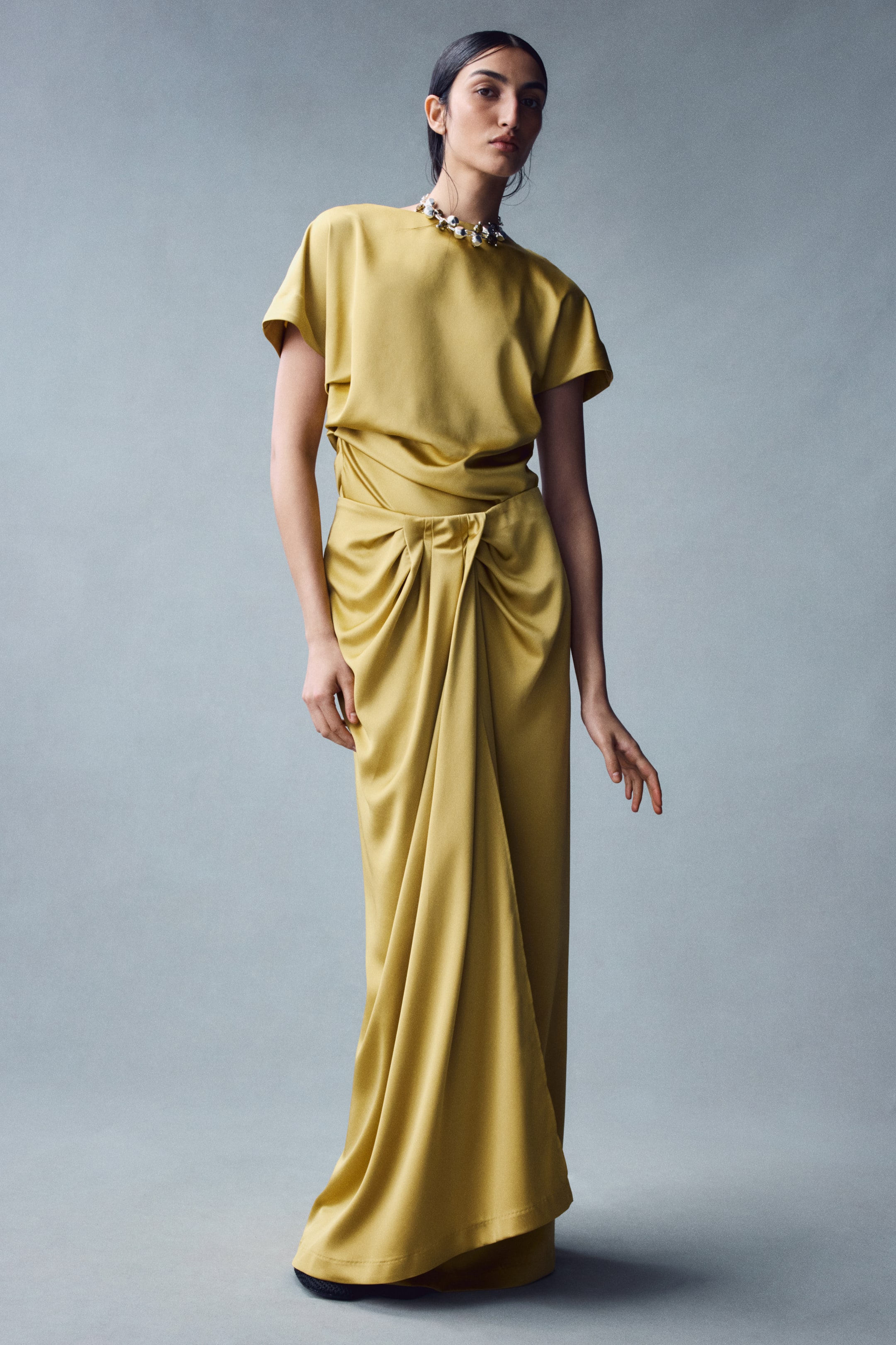 View larger image: The model is wearing a draped-detail satin skirt in a mustard-yellow shade, featuring a high waist and cascading gathers that create vertical folds down the front. The maxi-length skirt is styled with a matching short-sleeved satin top in the same colour.