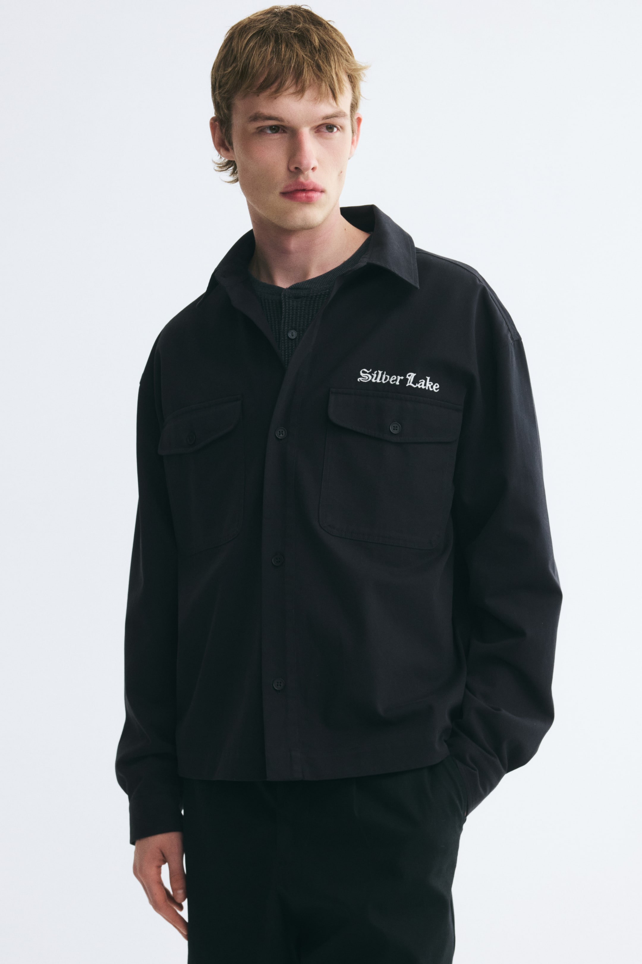 View larger image: The model is wearing a black cotton twill overshirt featuring a collar, button-down front, and buttoned flap chest pockets. The overshirt is layered over a dark grey textured shirt and features white embroidered text reading "Silver Lake" above the left chest pocket.
