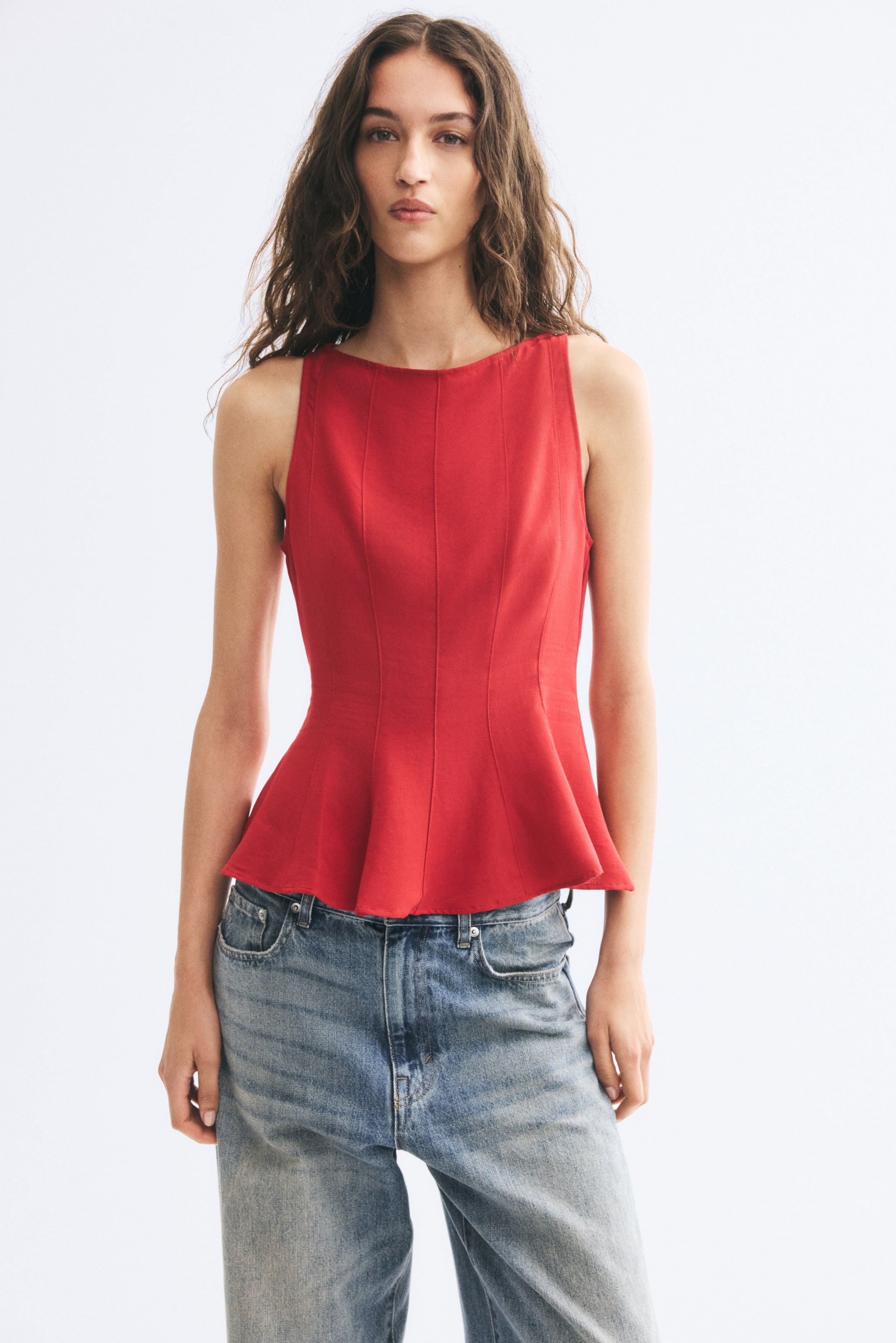 View larger image: A red, sleeveless peplum top featuring a boat neck and visible vertical seams down the front is worn by the model. The top flares at the waist and is paired with light-wash blue denim jeans.