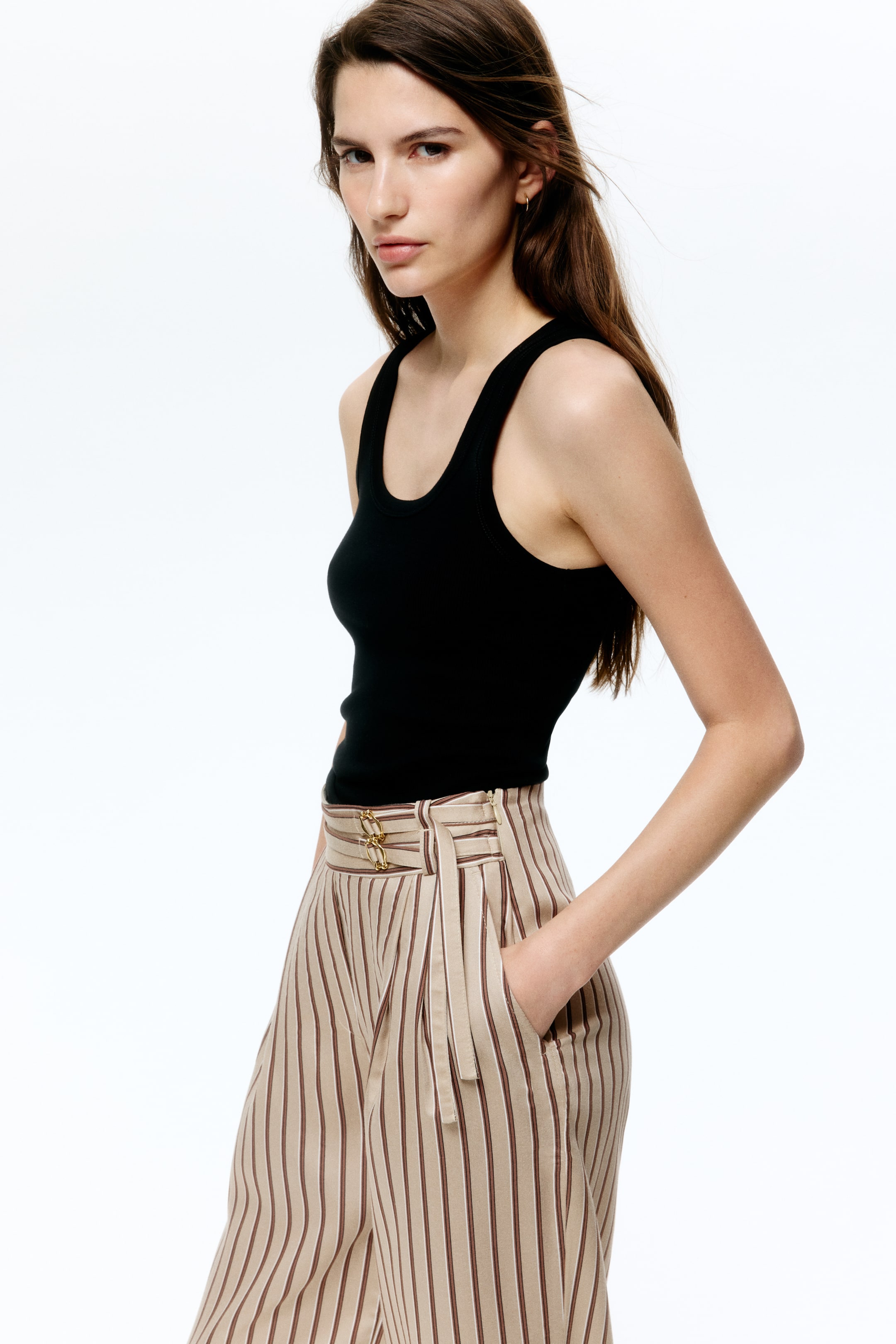 Visualizza immagine più grande: Pantaloni a gamba ampia e vita alta dalla vestibilità morbida, in fantasia a righe verticali beige e marroni, indossati dalla modella, con gamba larga, pieghe frontali, tasche laterali oblique e dettaglio decorativo a catena dorata sulla cintura. La modella ha una mano infilata in tasca e indossa un top a canotta nero a costine, infilato nei pantaloni.