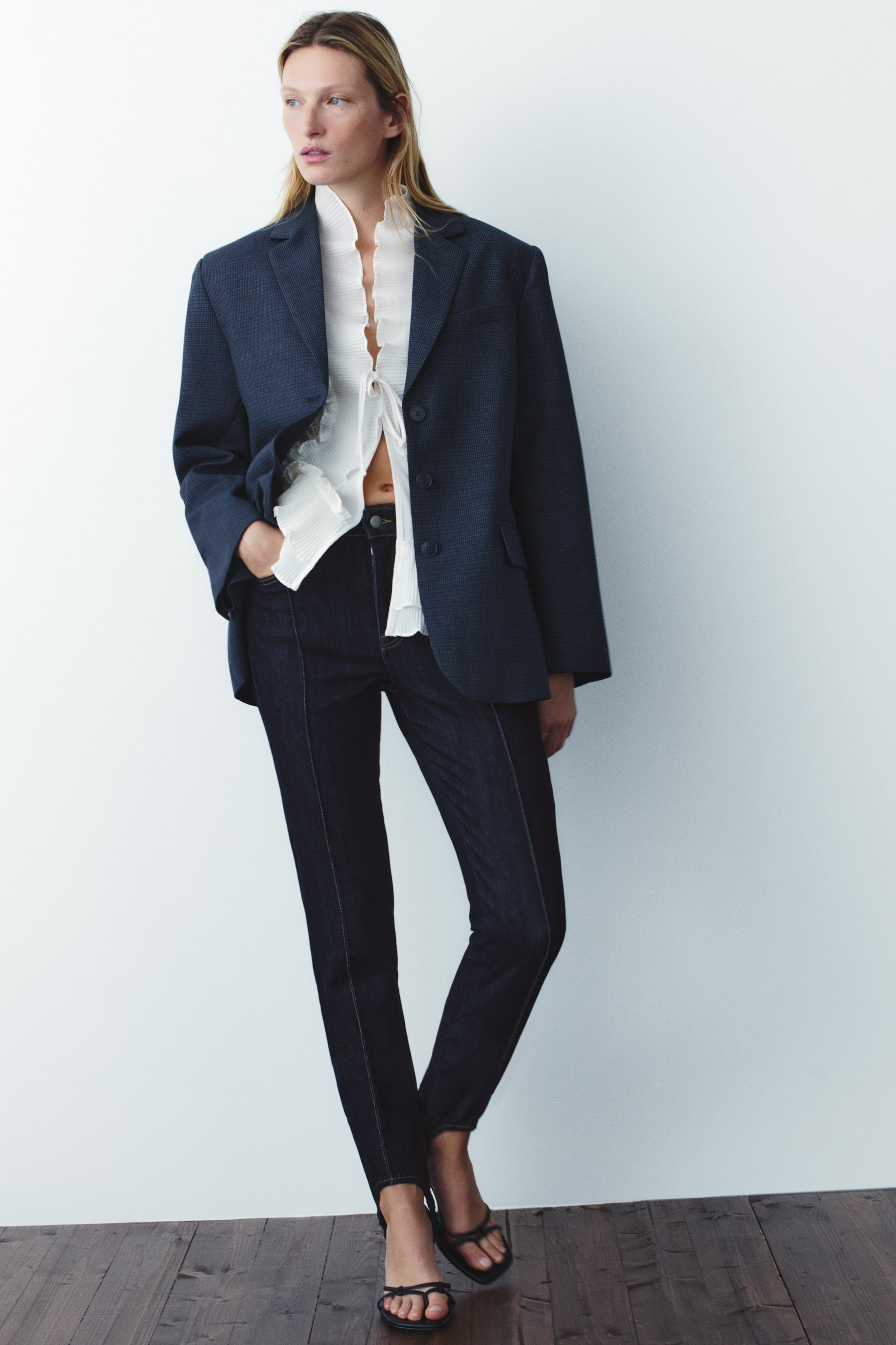 Visualizza l'immagine ingrandita: Jeans in denim scuro con staffa, caratterizzati da vita bassa, gambe slim con cucitura frontale visibile e cinturini sotto il piede, indossati dalla modella. La modella è in piedi con una mano infilata in una tasca frontale e abbina i jeans a una blusa bianca con volant, un blazer blu navy e sandali neri con cinturini.