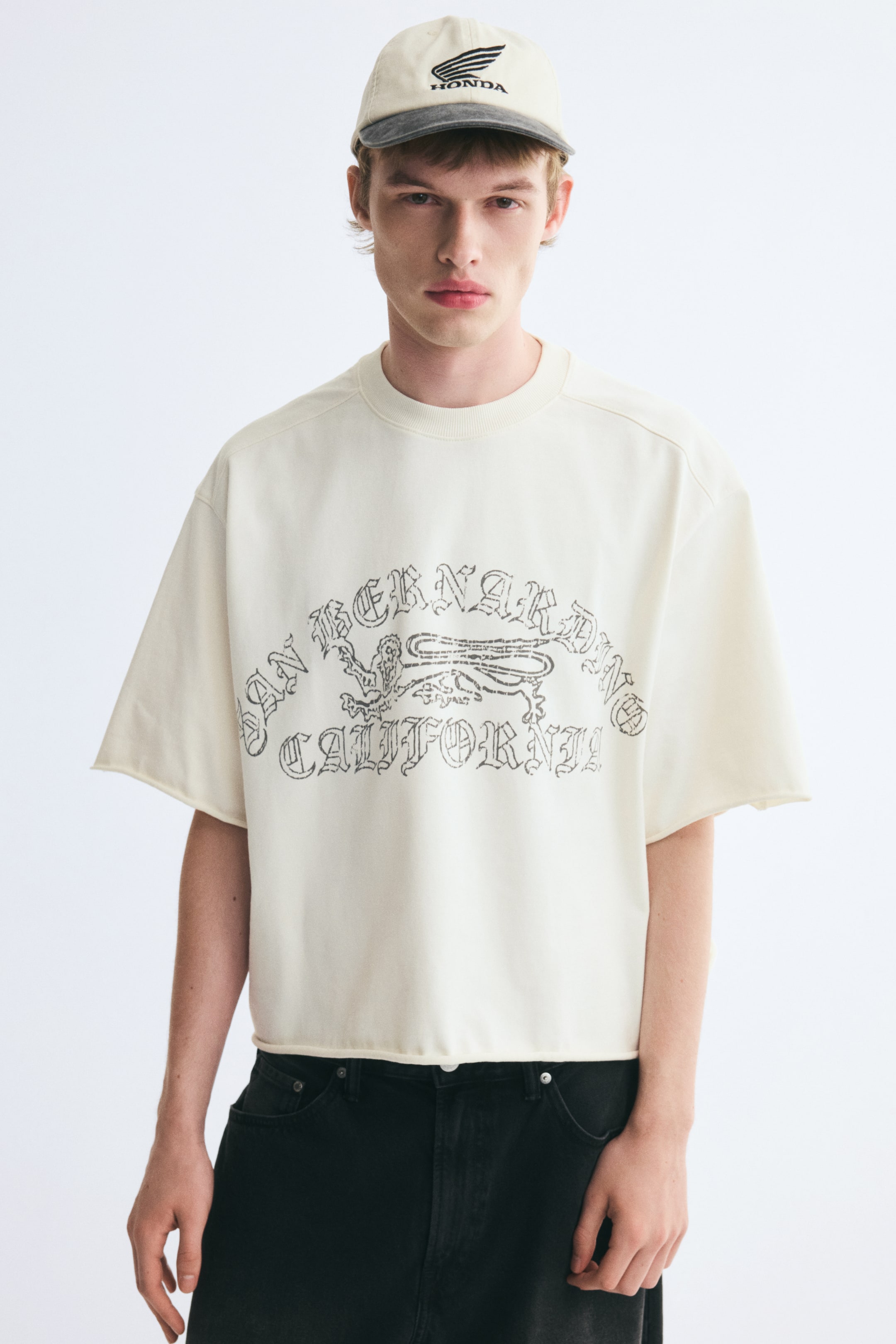 View larger image: The model is wearing an off-white, boxy-fit T-shirt featuring a printed graphic on the front. The oversized tee is designed with dropped shoulders, a ribbed crew neckline, and raw rolled edges at the cuffs and hem, styled with black jeans and a baseball cap.