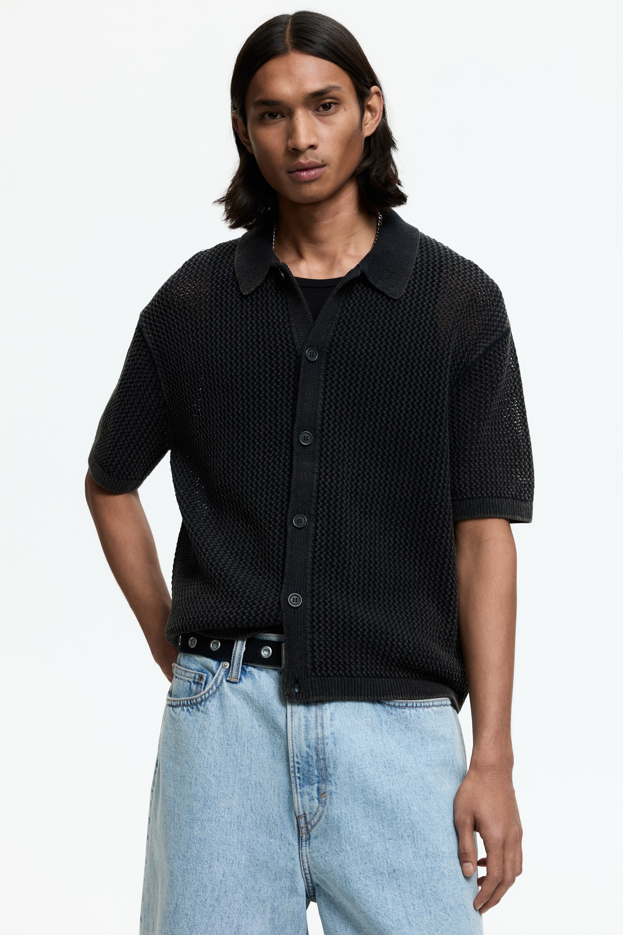 View larger image: The model is wearing a black short-sleeve hole-knit shirt with a collar, button-down front, and a straight hem. This relaxed-fit shirt is styled open over a black undershirt, with a few buttons fastened.