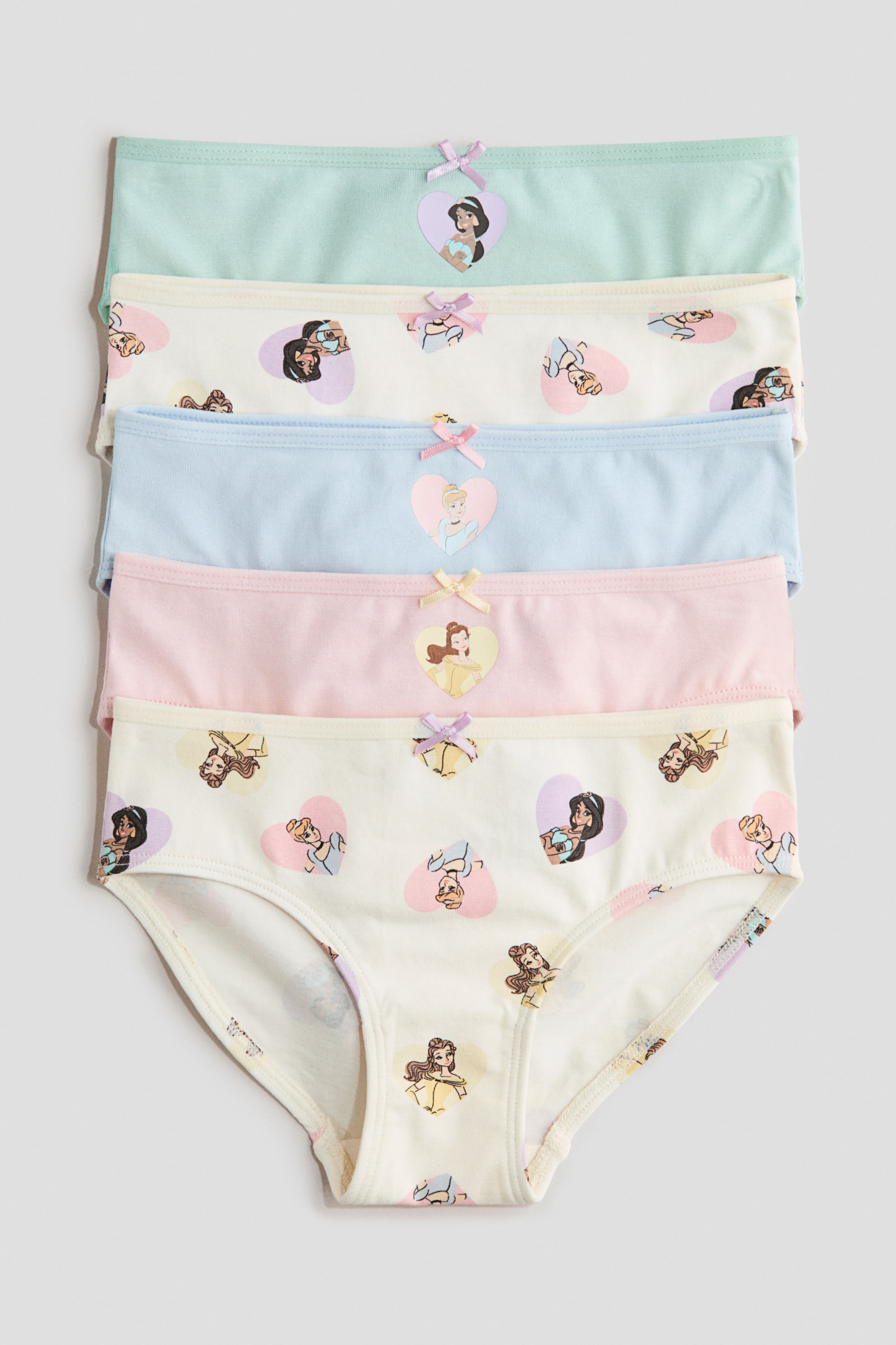 View larger image: Five pairs of children's briefs are neatly stacked, facing front. Three solid-colored pairs, in light green, light blue, and light pink, feature a heart-shaped character patch and a small bow on their elasticated waistbands. The two off-white pairs display an all-over pattern of multiple heart-shaped character patches and small bows on their elasticated waistbands.