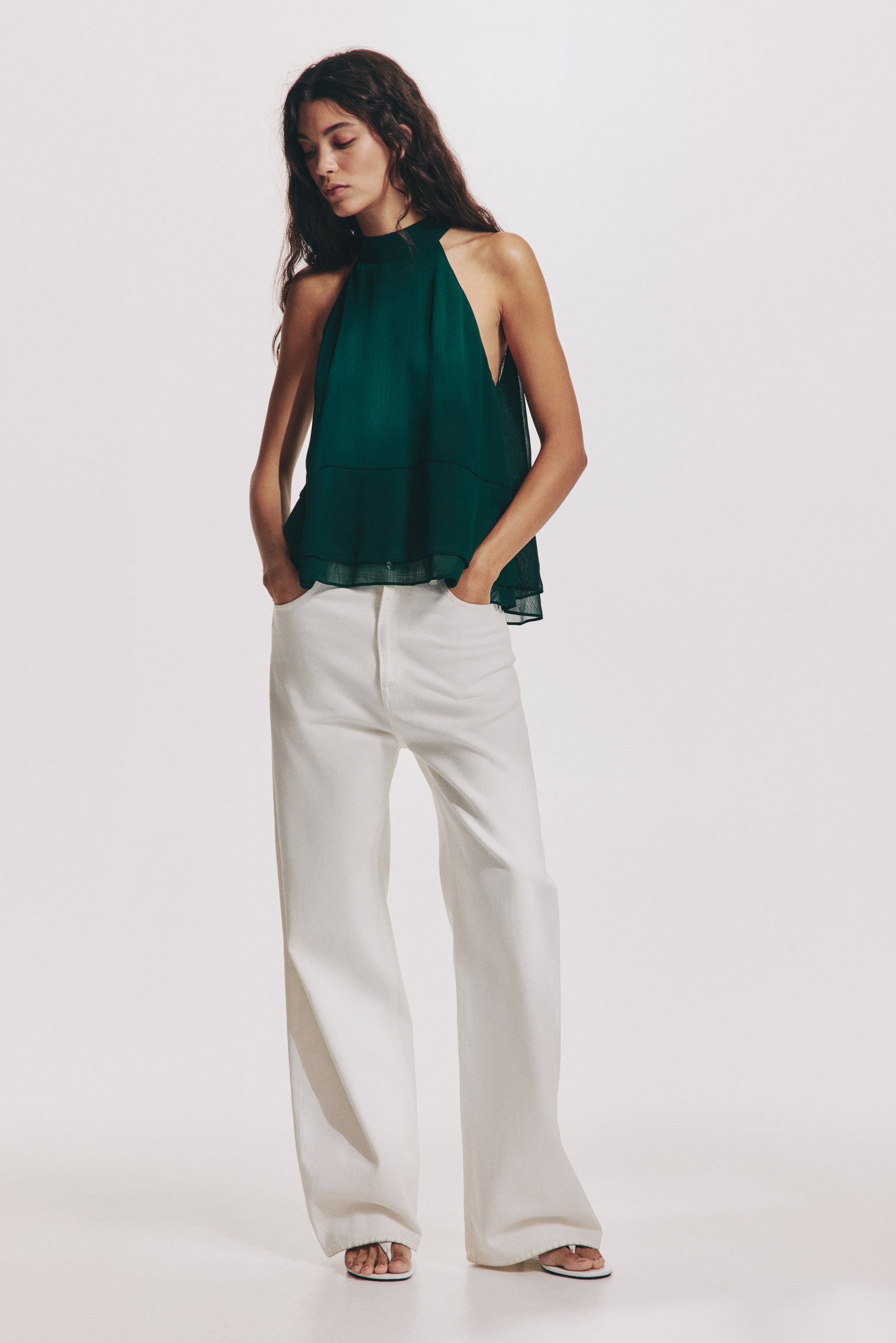 View larger image: A dark green halterneck top, crafted from a sheer, crinkled weave, is being worn by the model. The top features a narrow cut at the top with wide ties, a V-shaped opening at the back, and a wide, double-layered flounce at the hem.
