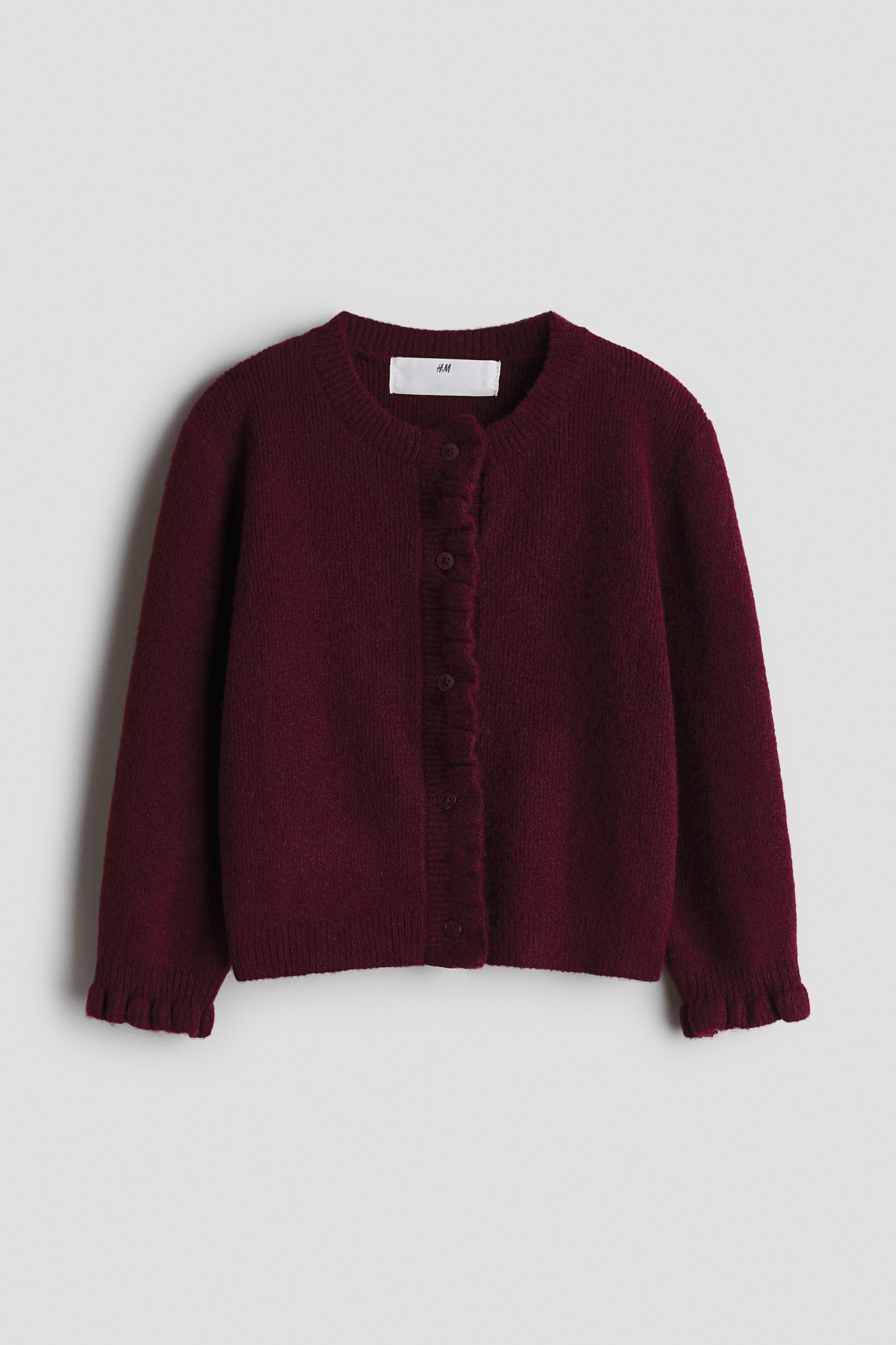 View larger image: A burgundy cardigan is shown from the front, featuring a round neck and button-up fastening. It has a ruffled placket and ruffled cuffs, with small round buttons matching the cardigan’s colour.