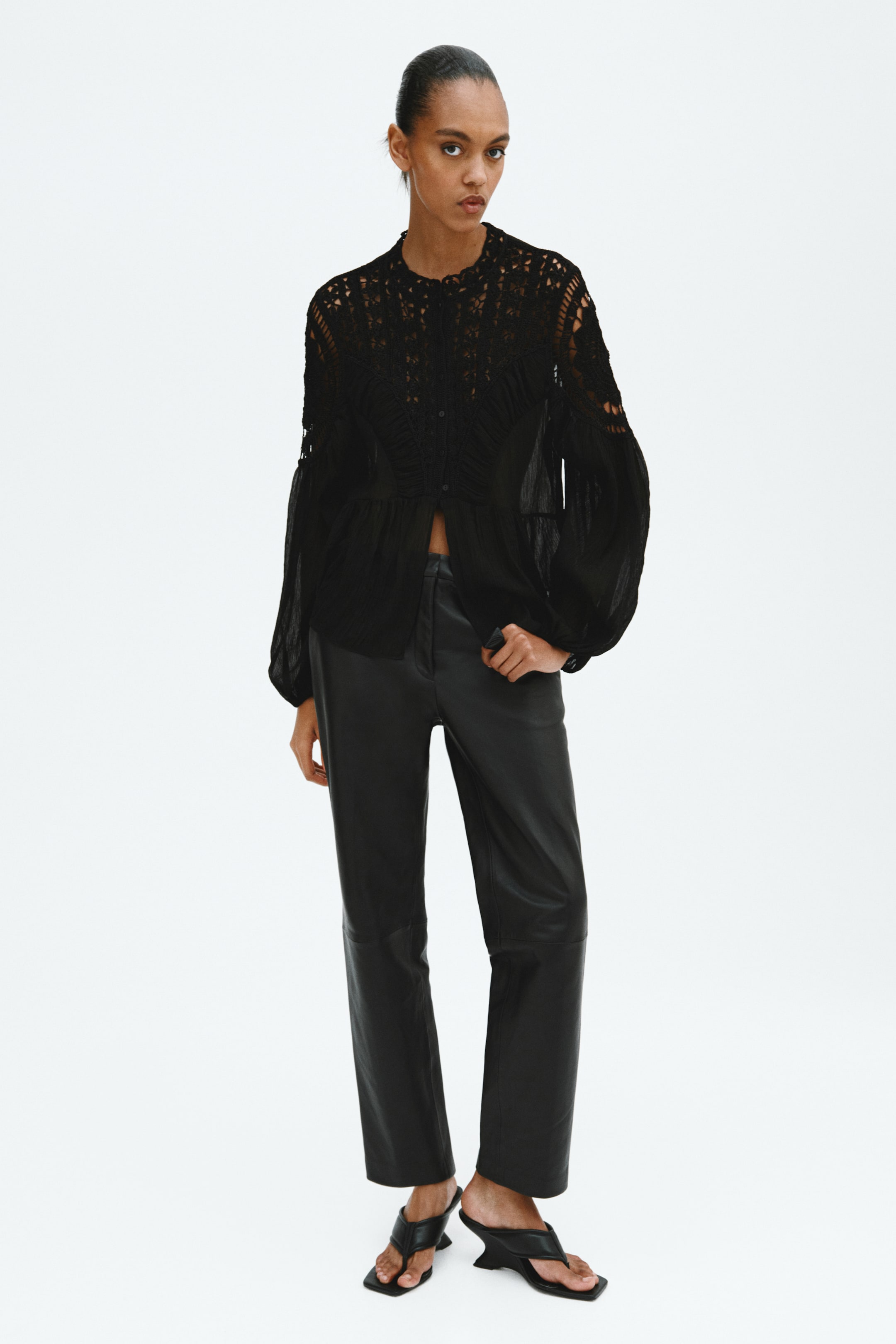 View larger image: A black long-sleeved blouse featuring intricate crochet detailing on the upper chest and shoulders, a ruffled collar, and gathered sleeves is paired with black straight-leg leather trousers displaying a front seam and black square-toed thong sandals with a sculptural heel.