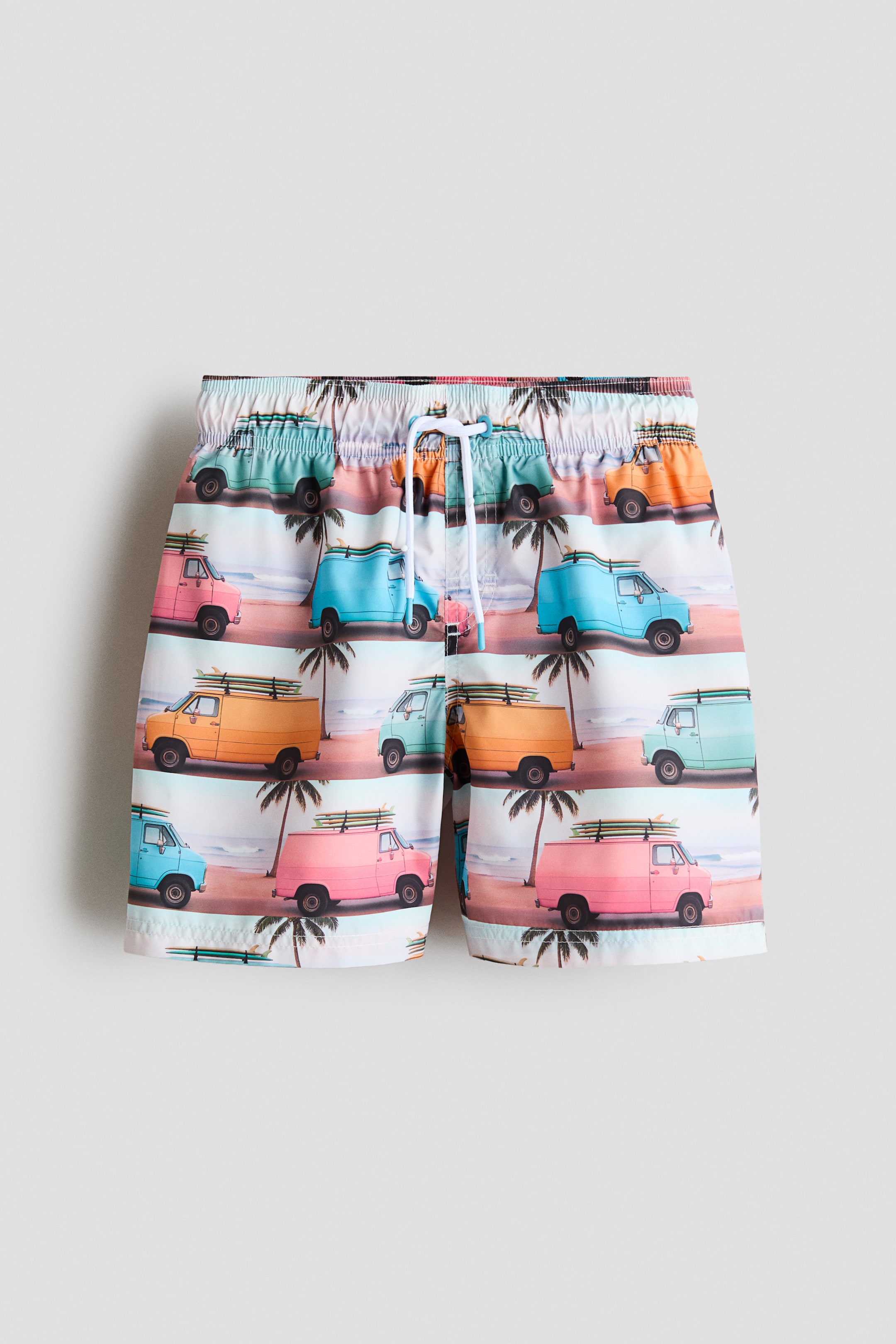 Visa större bild: Tryckta badshorts visas framifrån, med en elastisk midja med vit dragsko och en sydd fejkad gylf. Shortsen har ett återkommande mönster av färgglada vintagebussar med surfbrädor, mot en bakgrund av strand och palmer.