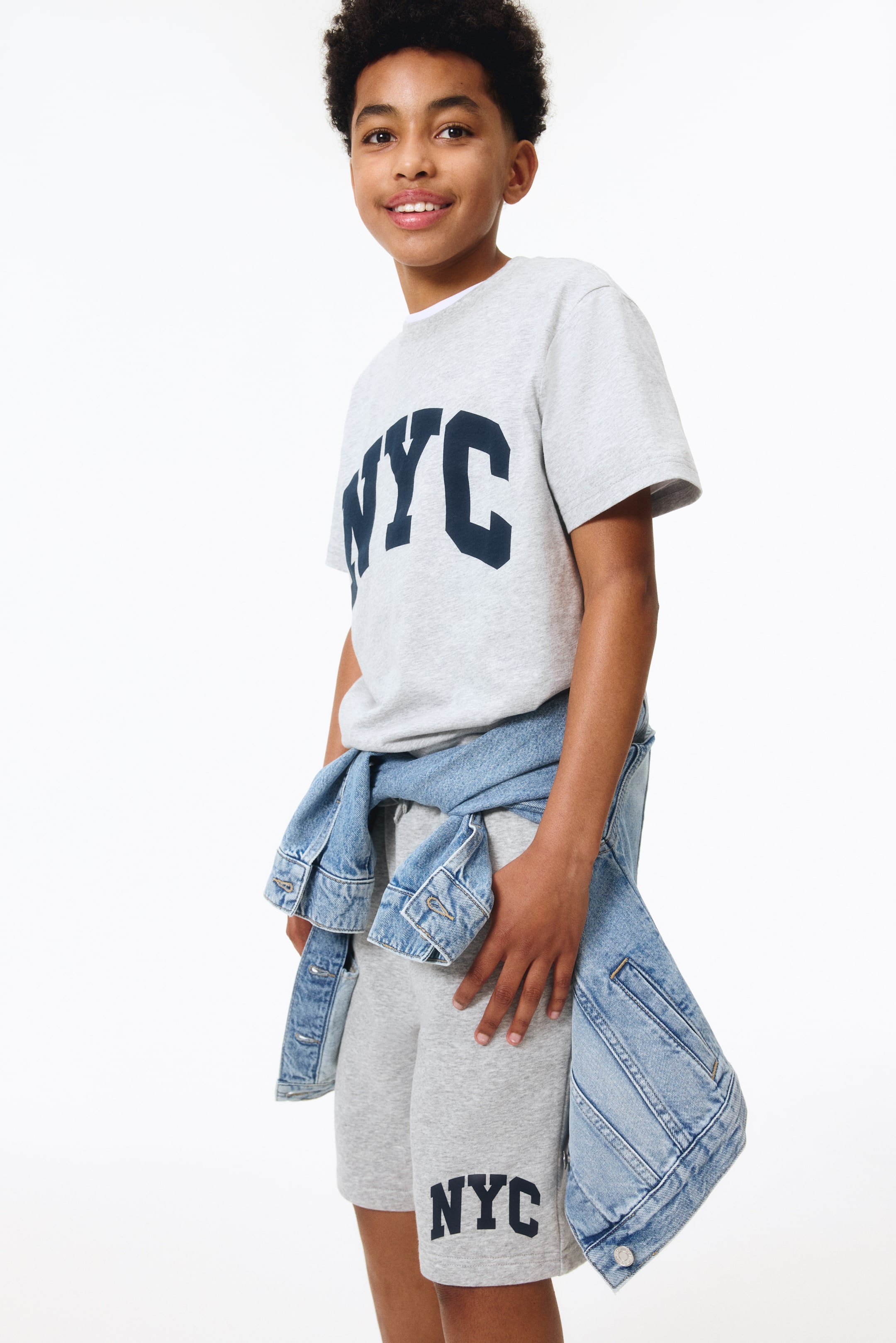 Ver imagem maior: T-shirt clássico apresenta uma vista frontal, construído em tecido jersey de algodão cinza claro estampado. Exibe uma gola redonda canelada, mangas curtas e 'NYC' em letras grandes e azuis escuras no peito.