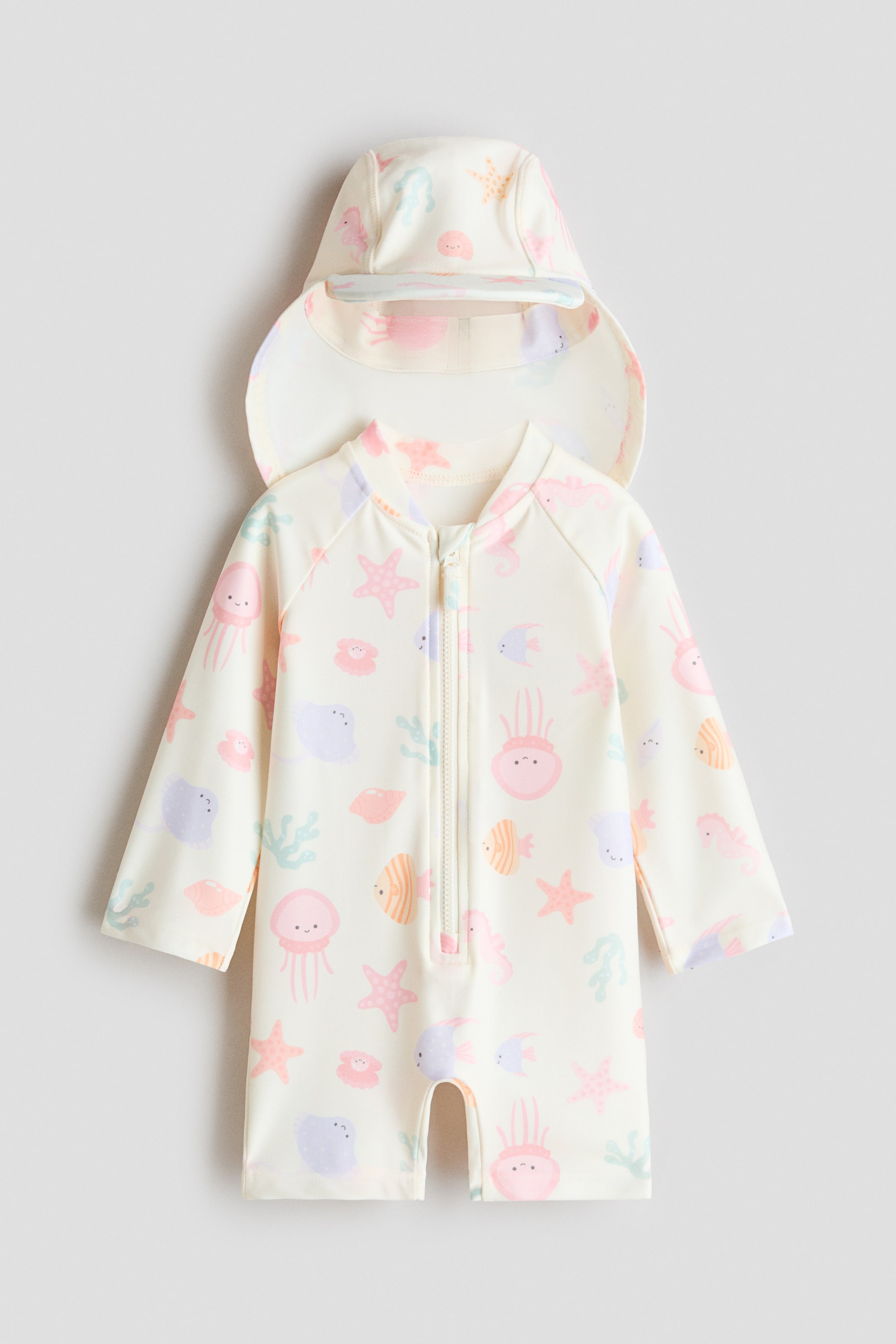 Ampliar la imagen: Un conjunto de protección solar color crema, que incluye un traje de baño y una gorra a juego, presenta un estampado colorido de criaturas marinas. El traje de baño, orientado hacia el frente, muestra un pequeño cuello alto, mangas largas raglán, piernas cortas y un cierre frontal. La gorra coordinada presenta una visera en la parte delantera y una solapa que cubre la parte trasera del cuello.