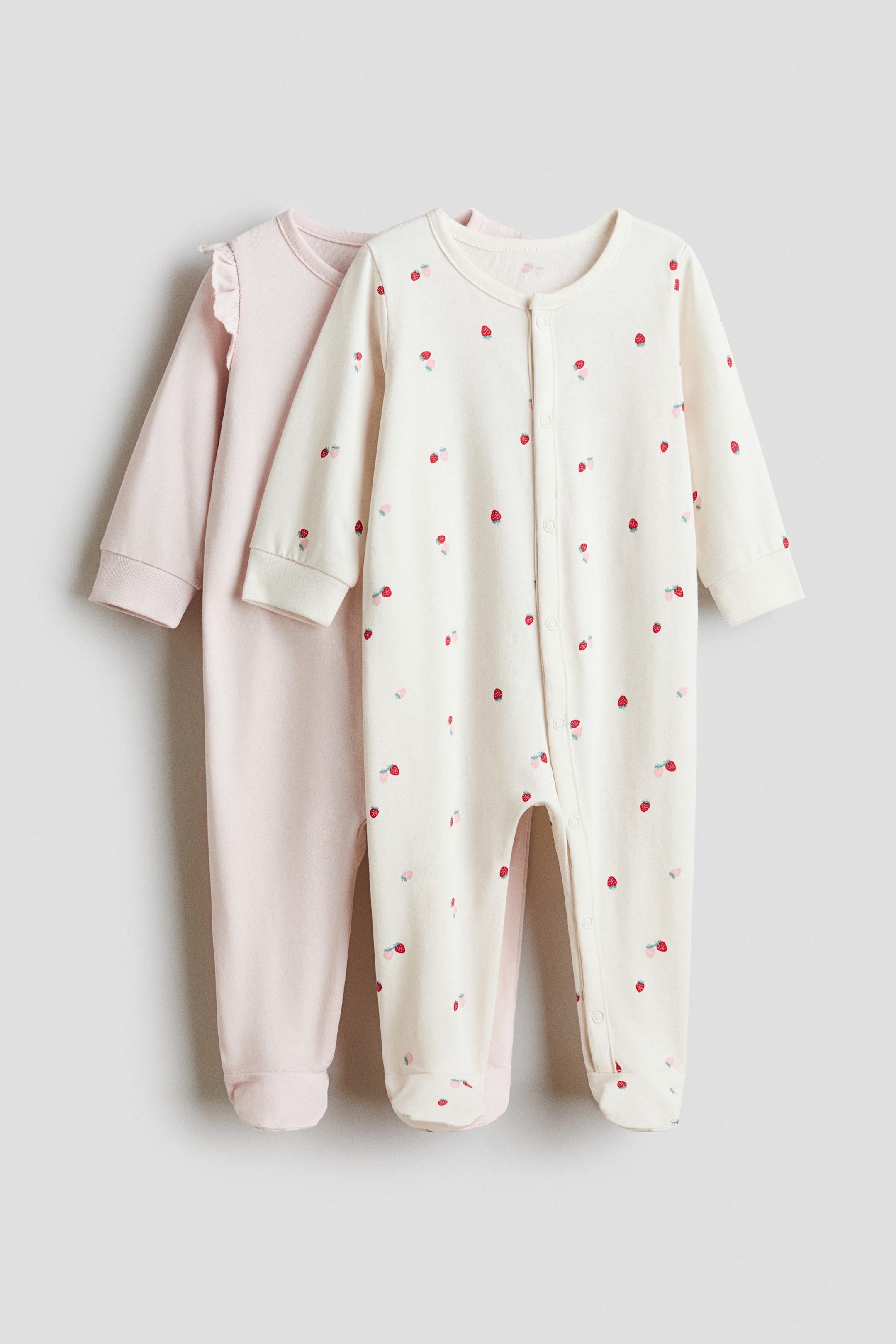 View larger image: A 2-pack of cotton sleepsuits is presenting. The FRONT cream sleepsuit is visible, showing a scattered red and pink strawberry print, long sleeves with cuffs, a round neckline, and a snap-button closure that is running down its front and inner leg, with integrated footies, while a second light pink sleepsuit is positioned behind, also showing long sleeves with ruffled shoulder details and integrated footies.