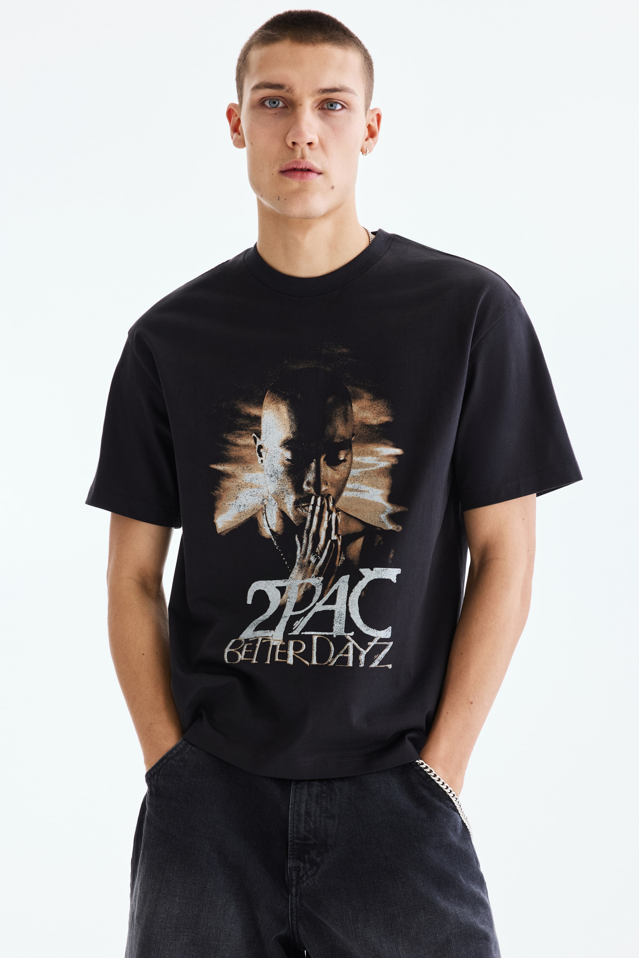 Größeres Bild anzeigen: Ein schwarzes T-Shirt aus weichem, bedrucktem mittelschwerem Baumwolljersey, das einen Grafikdruck von 2Pac mit gefalteten Händen und dem Text „2PAC BETTER DAYZ“ auf der Vorderseite zeigt, wird vom Model getragen. Es hat überschnittene Schultern, einen runden Halsausschnitt mit schmaler Einfassung und einen lockeren Schnitt.
