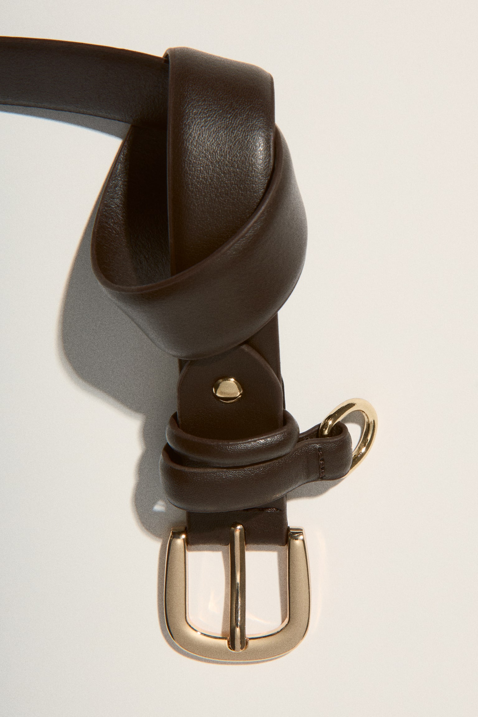 A narrow, dark brown belt is lying flat, featuring a gold-toned rectangular buckle with a single prong. A matching dark brown keeper with a small gold-toned D-ring is also visible near the buckle. - 1