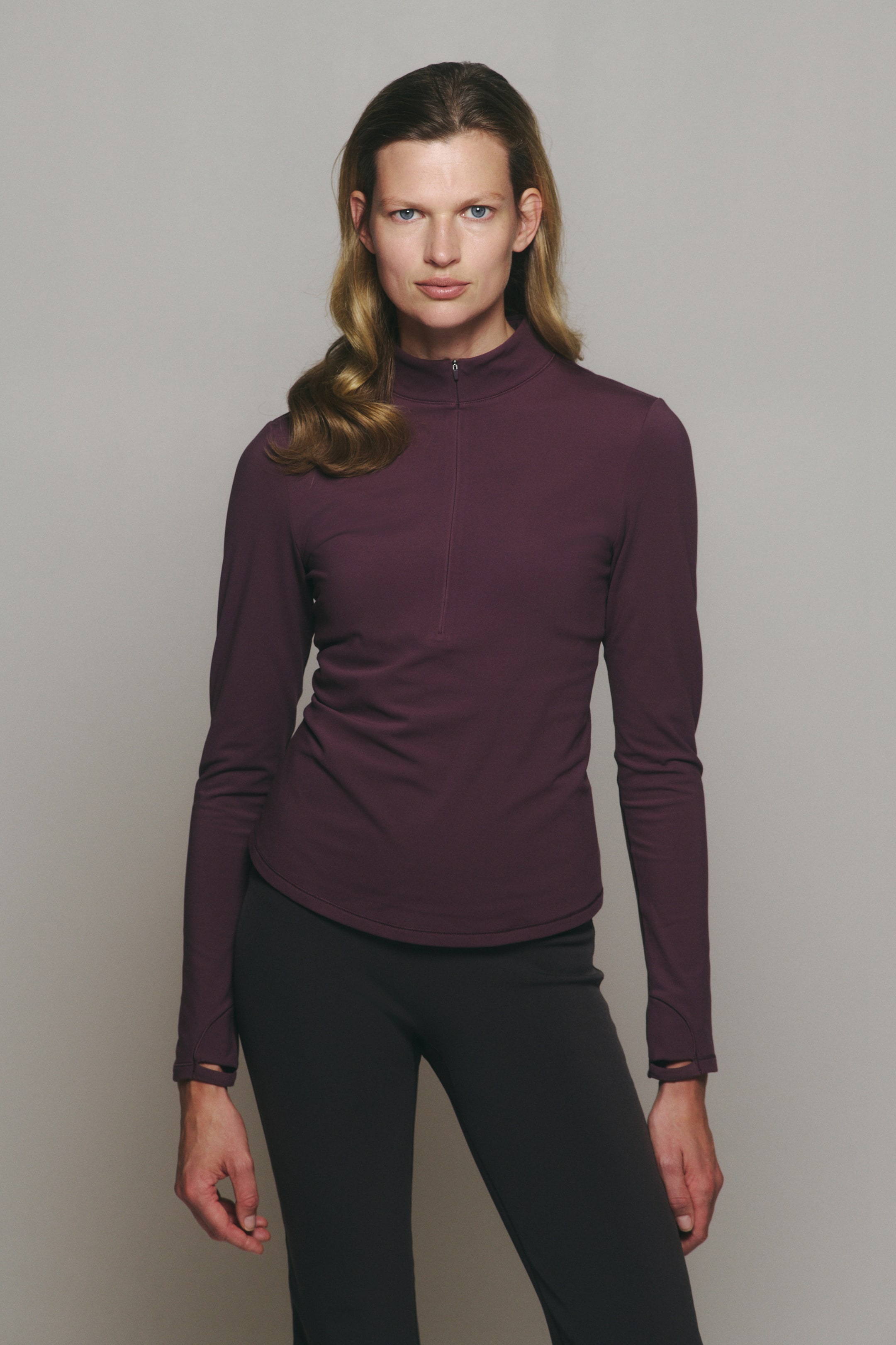 Agrandir l'image: Un haut à manches longues violet foncé avec fermeture éclair quart de longueur, doté d’un col montant et de poignets avec passe-pouces, est porté par le mannequin. Le haut est associé à un pantalon de sport gris foncé.
