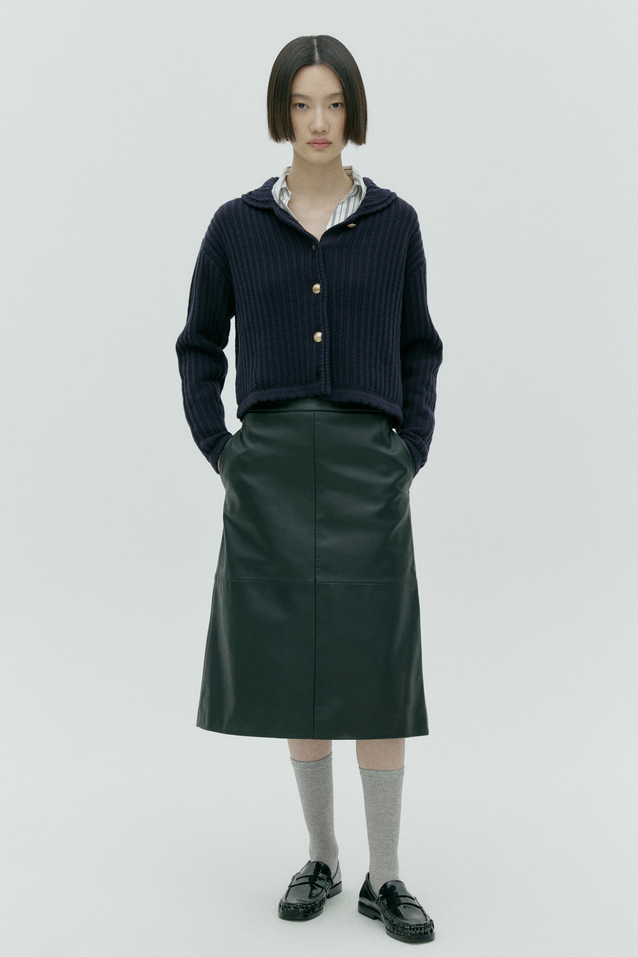 拡大画像を表示: ネイビーブルーのリブニットカーディガンは、クロップド丈とスプレッドカラーで仕立てられています。前立てにはゴールドボタンが配され、ストライプシャツにレイヤードされています。