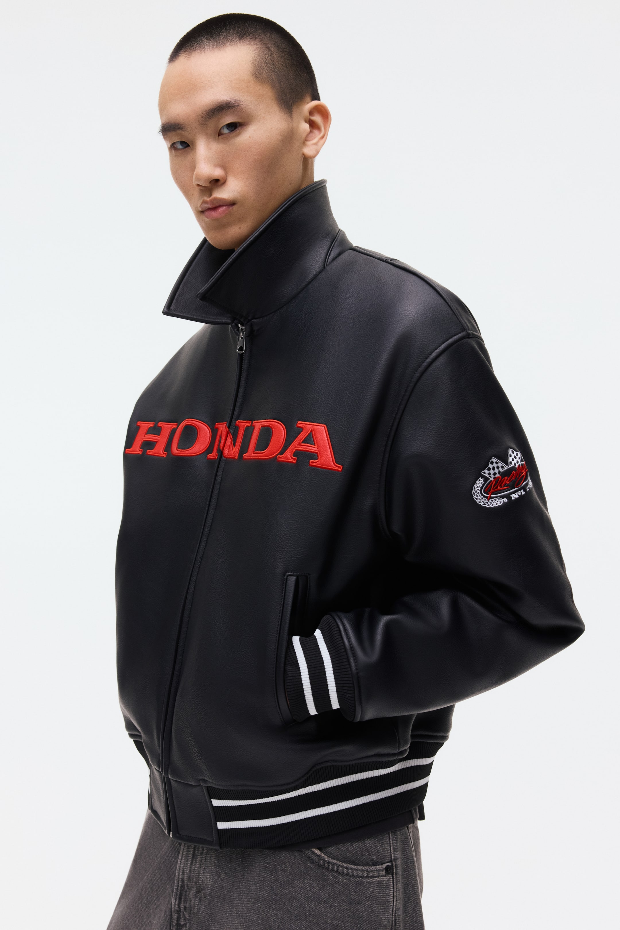 Voir une image plus grande: Une veste en similicuir noir est présentée sous un léger angle, mettant en valeur son col montant, sa fermeture éclair complète à l'avant et son côté gauche. Le lettrage "HONDA" en grandes lettres rouges est brodé sur la poitrine, avec un écusson de drapeau à damier sur la manche gauche. La veste est également dotée de poignets côtelés et d'un ourlet côtelé, tous deux détaillés avec deux bandes blanches, et comprend des poches passepoilées en biais.