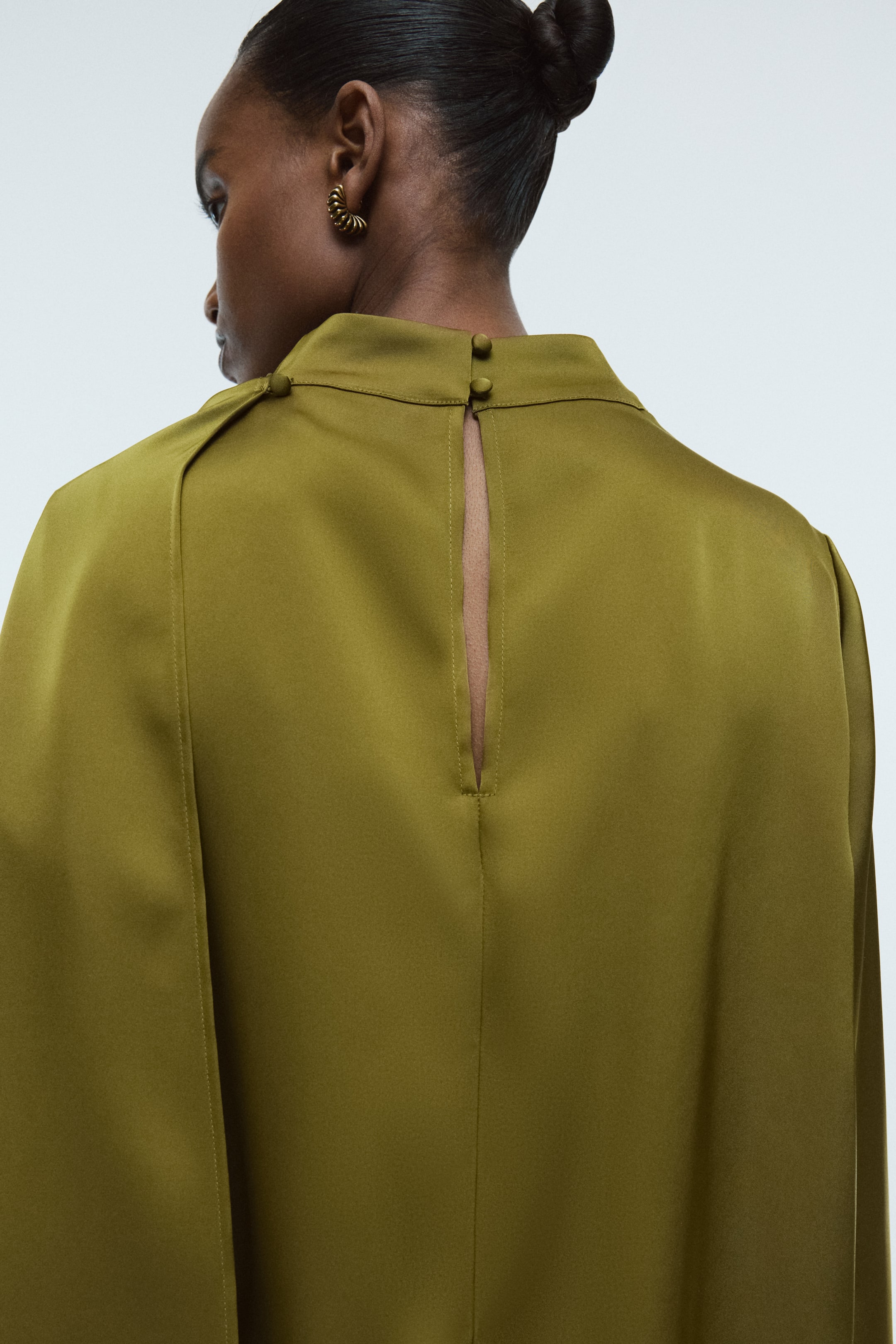 View larger image: A blouse in lustrous olive green satin is being worn by the model, featuring a stand collar and a keyhole opening at the back of the neck, fastened with two covered buttons.