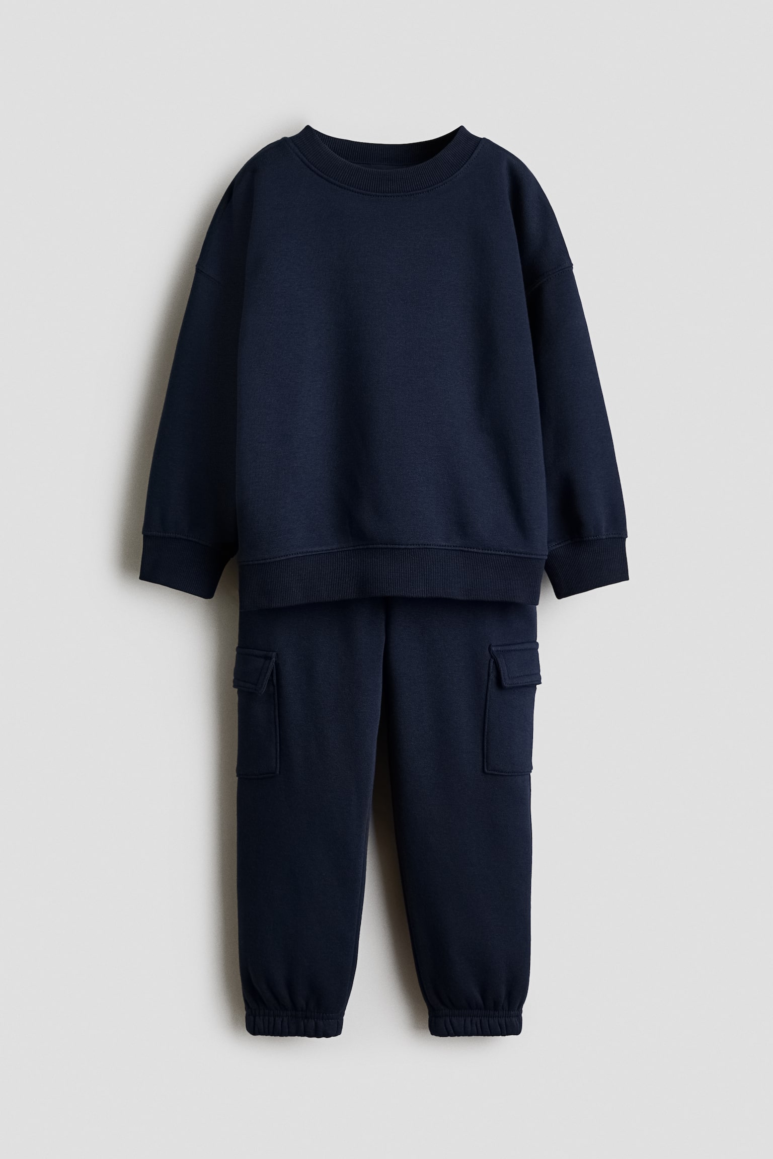 Un ensemble deux piÃ¨ces en bleu marine composÃ© d'un sweat-shirt et d'un pantalon est prÃ©sentÃ© de face, avec un sweat-shirt Ã  manches longues dotÃ© d'un col rond cÃ´telÃ©, d'Ã©paules tombantes, de poignets cÃ´telÃ©s et d'un ourlet cÃ´telÃ©. Le pantalon jogger assorti affiche des poches cargo latÃ©rales Ã  rabat ainsi que des chevilles resserrÃ©es par des poignets Ã©lastiquÃ©s.
