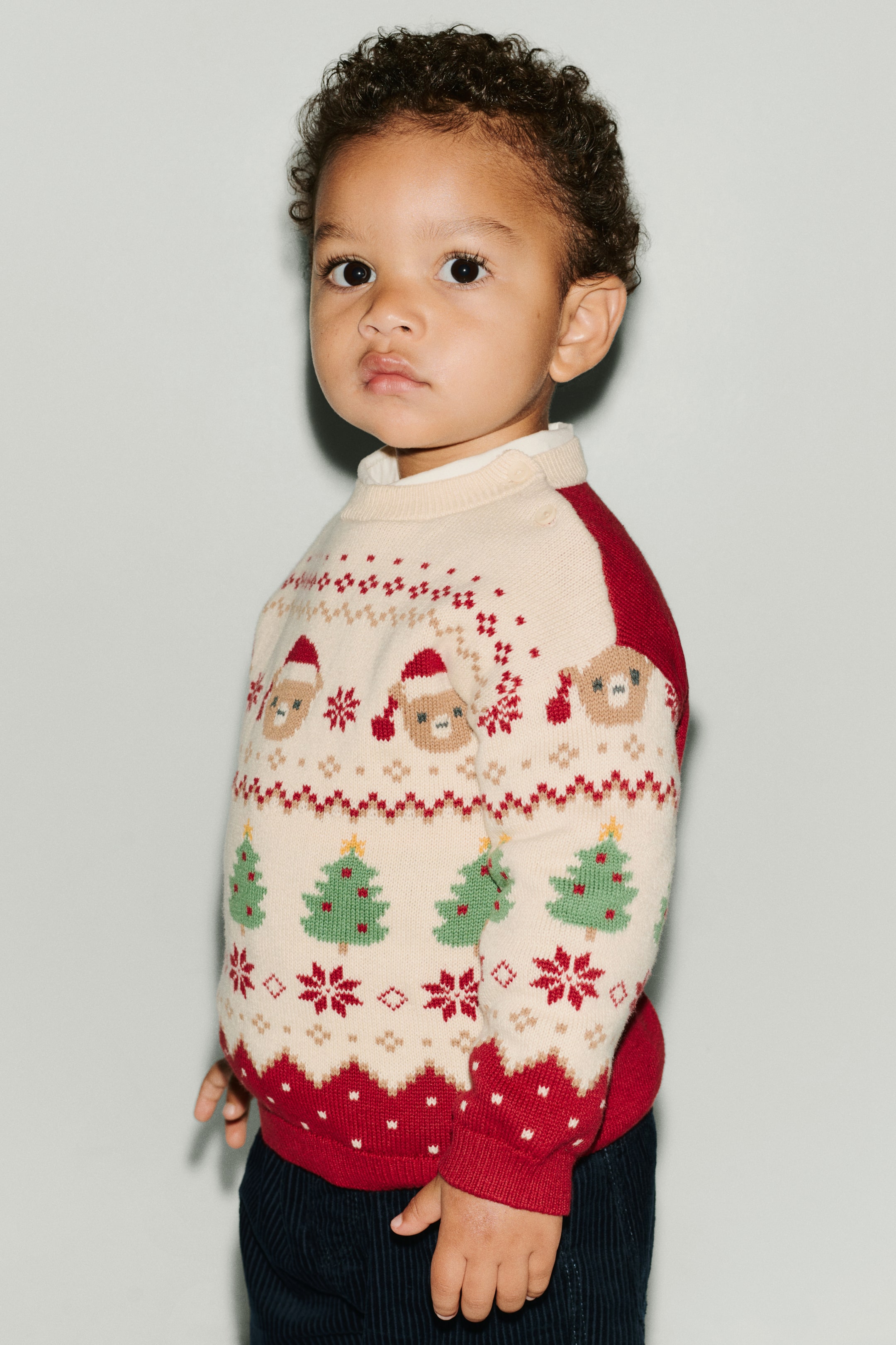 View larger image: This jacquard-knit sweater, shown from a three-quarter front angle, features a cream-coloured front panel patterned with festive motifs of trees, snowflakes, and holiday characters, alongside contrasting red sleeves and back. The sweater features a round, rib-trimmed cream neckline with button closures on one shoulder, dropped shoulders, and red ribbed cuffs and hem.