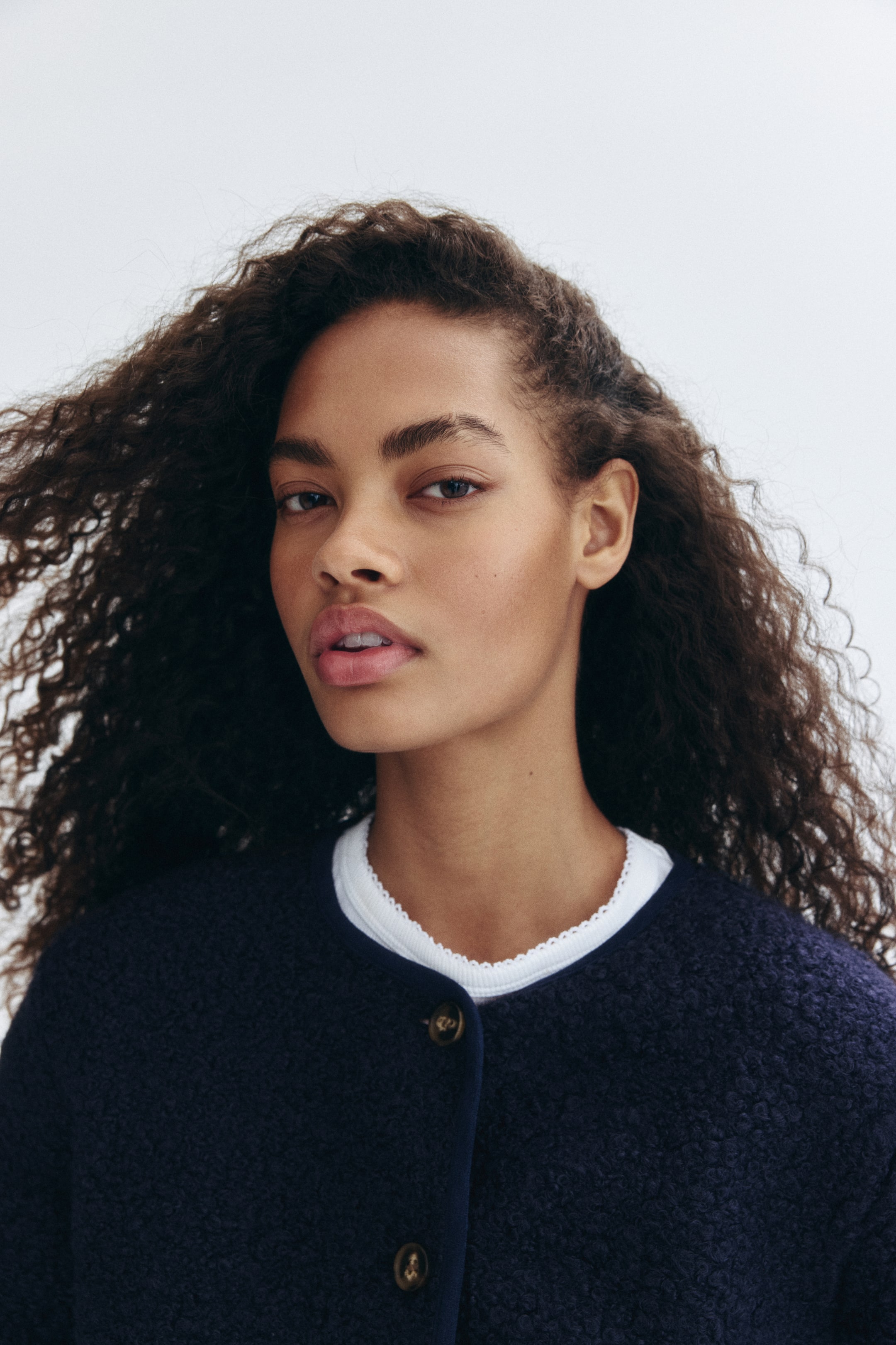 View larger image: A navy blue textured cardigan is being worn by the model, featuring a round neckline and two visible brown buttons on its front placket. A white top with a decorative scalloped trim is layering beneath the cardigan.