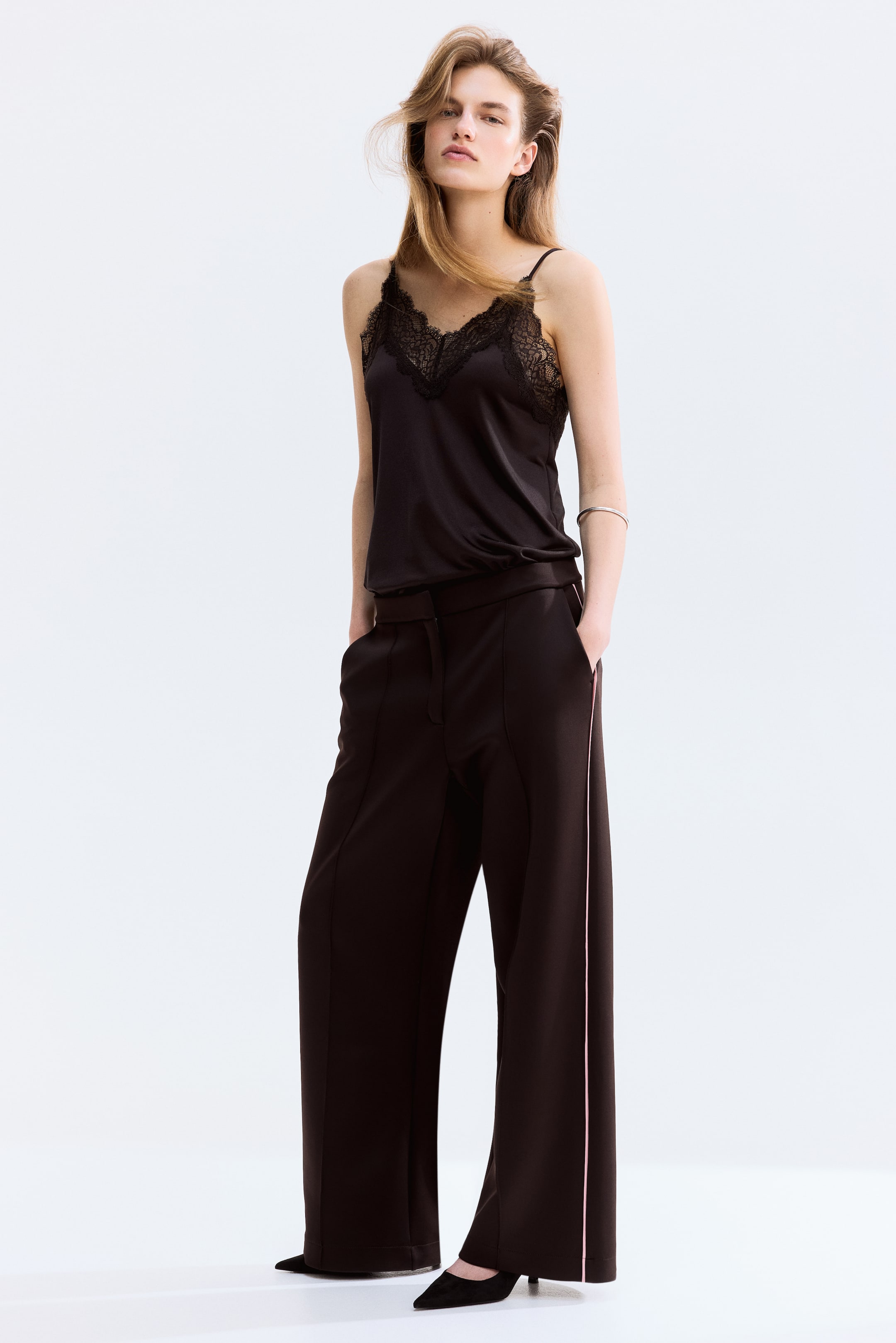 Visualizza immagine più grande: Un top in raso nero con scollo a V e rifiniture in pizzo nero viene presentato, abbinato a pantaloni neri a gamba larga con una striscia laterale rosa. Il top è infilato nei pantaloni.