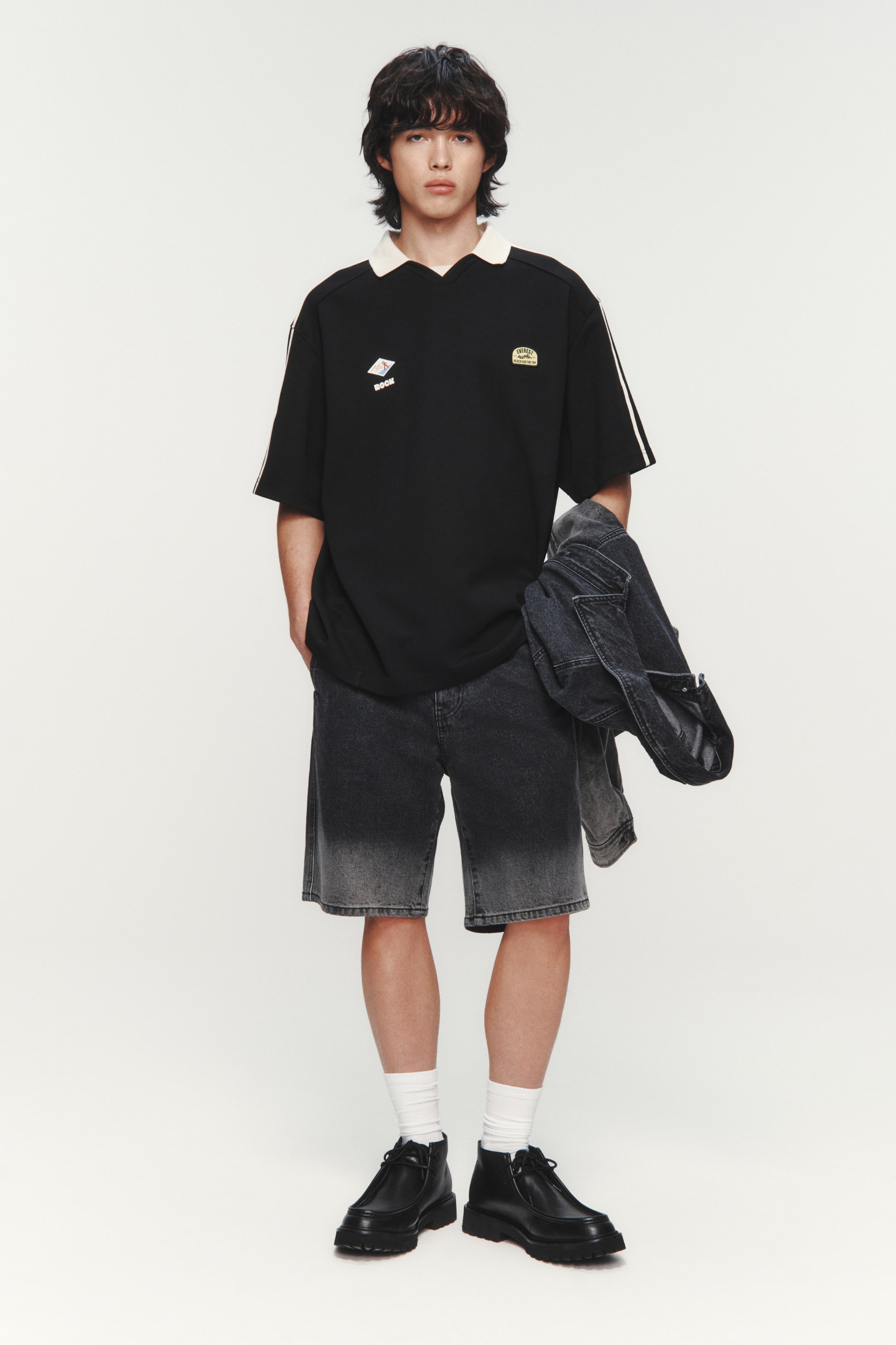 View larger image: A black polo shirt with an off-white collar, featuring two white stripes on each short sleeve and two embroidered patches on the left chest. Paired with dark grey denim shorts that fade to a lighter grey at the hem, and black ankle boots with lugged soles.